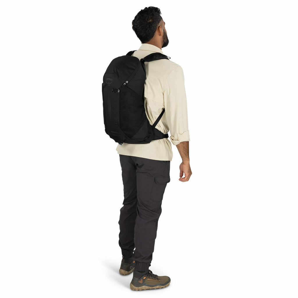 The image features a man standing against a plain white background, turned slightly away from the camera, showcasing a backpack. He is wearing an off-white long-sleeve shirt, dark grey pants, and brown hiking boots with a tan sole. The backpack is black a