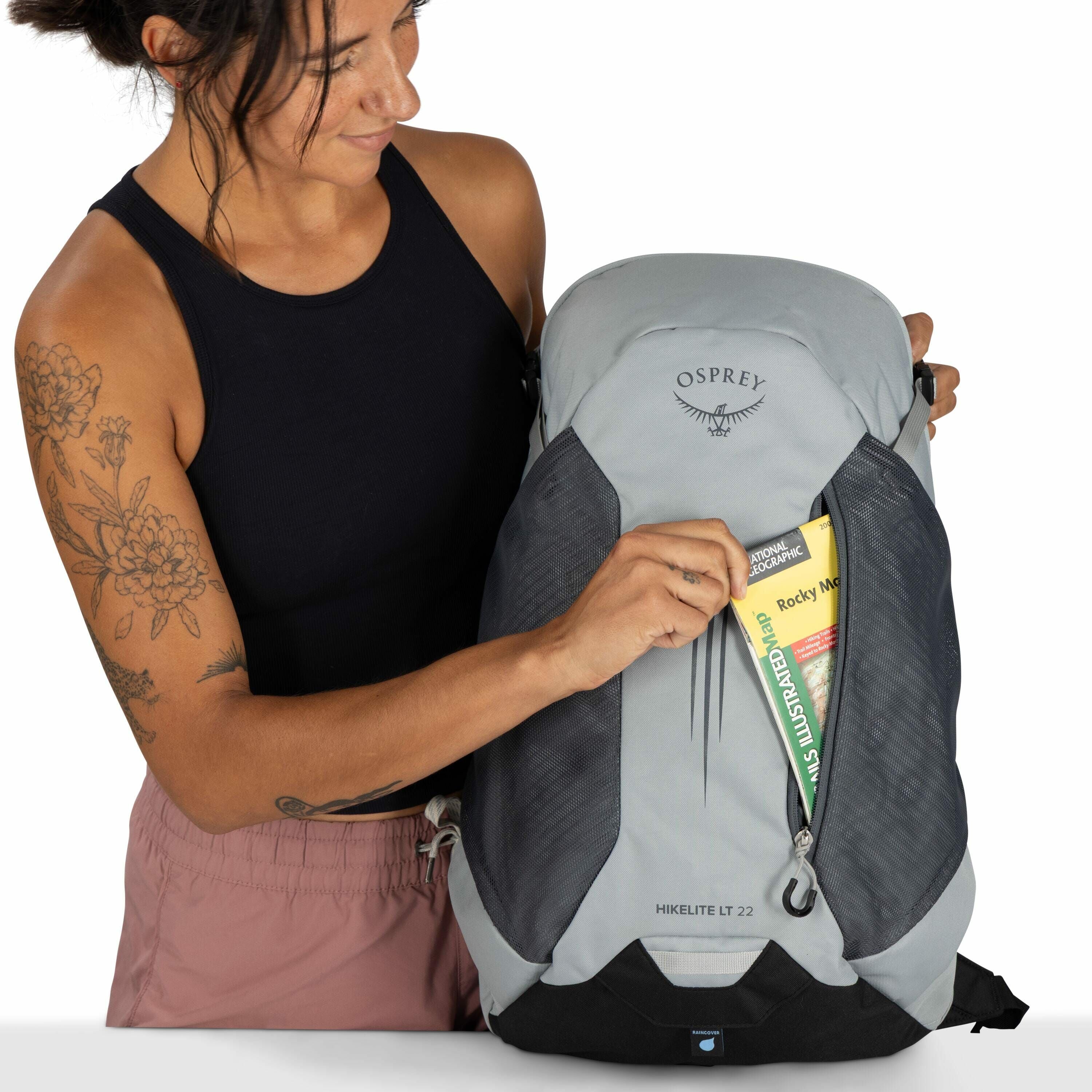 The image features a person holding a light gray Osprey Hikelite LT 22 backpack. The individual is wearing a black tank top and pink shorts, with visible floral tattoos on their left arm. They are placing a National Geographic map titled "Rocky Mountain"