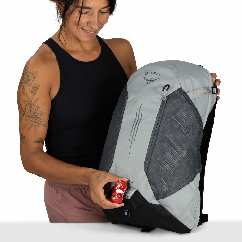 The image features a person holding an Osprey backpack with a light grey and black color scheme. The person is wearing a black sleeveless top and pinkish-brown shorts, and they have detailed floral tattoos on their left arm. They are holding a red object,