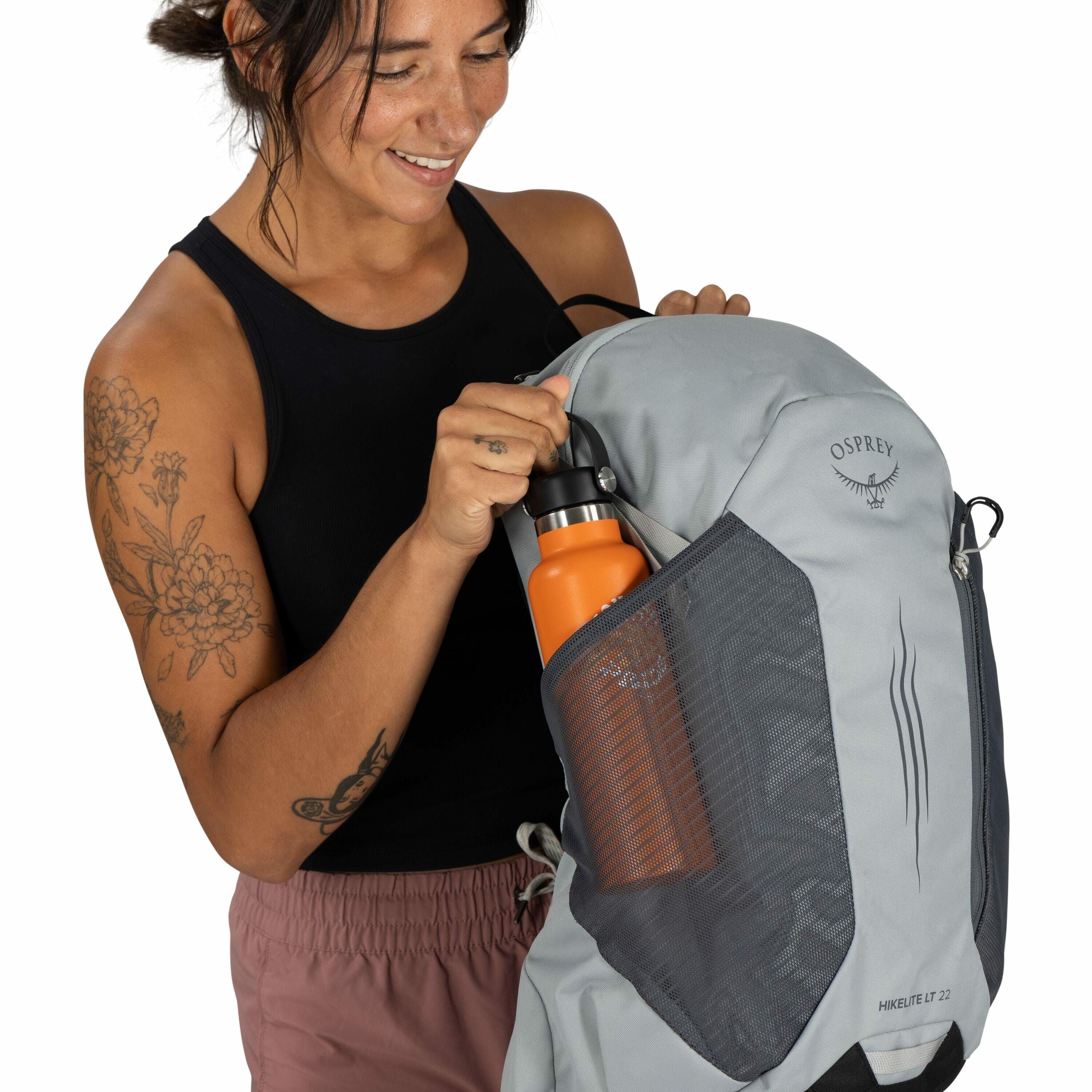 The image shows a person dressed in a black sleeveless top and rose-colored pants, holding a gray backpack made by Osprey. The backpack has mesh side pockets, and one of them contains an orange water bottle with a black lid. The backpack is marked with th