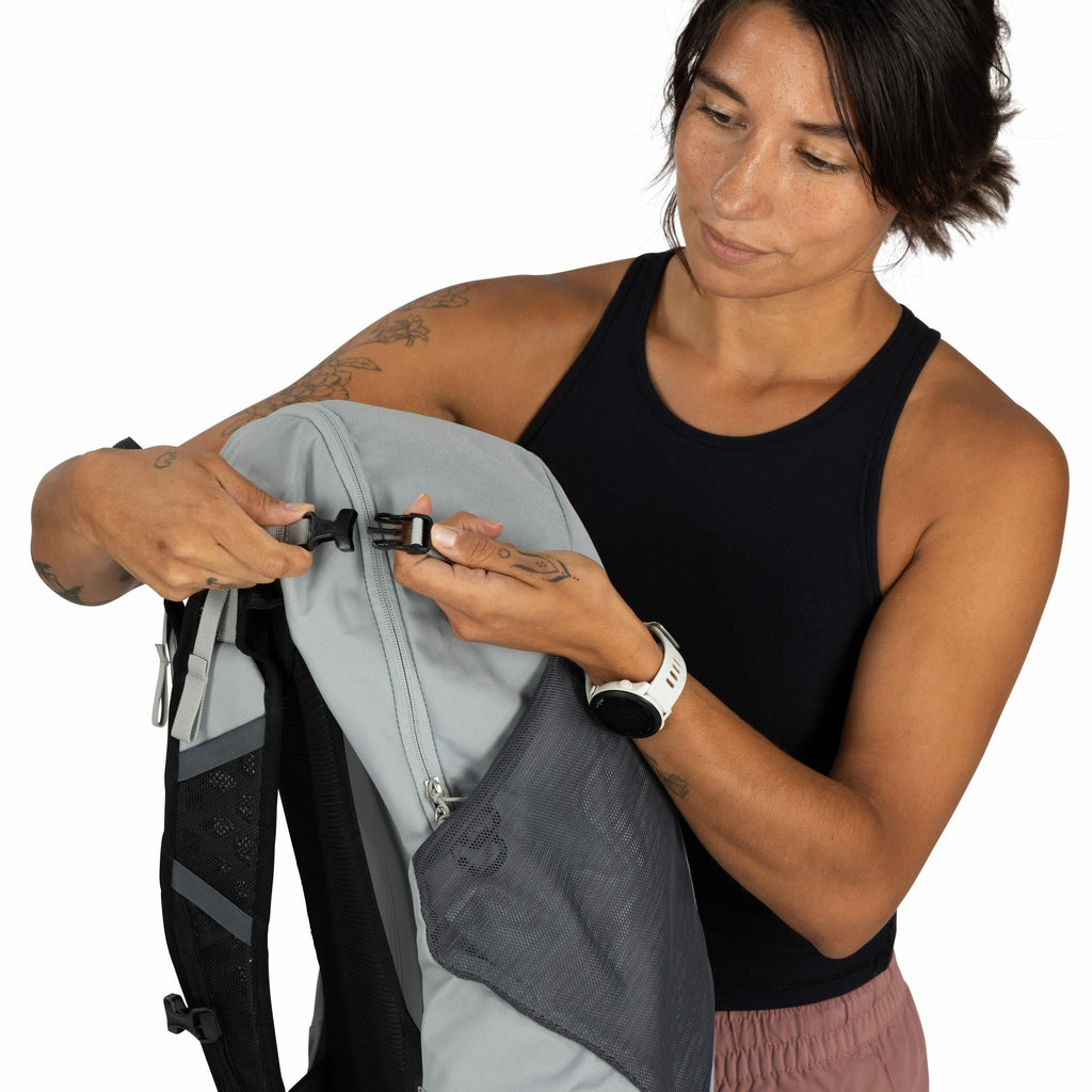 The image features a person holding a gray and black backpack, demonstrating the clipping mechanism. The person is wearing a black tank top and pinkish-brown pants, accessorized with a white smartwatch on their left wrist. Their arms are tattooed with var