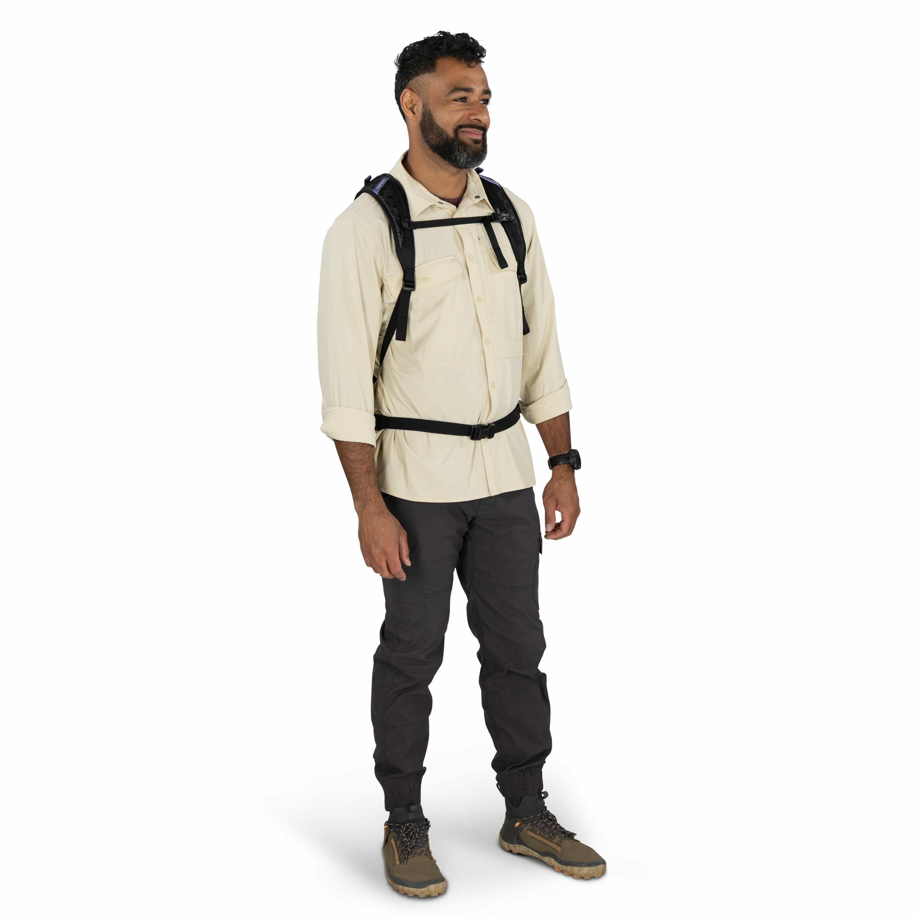 The image shows a man standing against a plain white background, dressed in outdoor hiking attire. He is wearing a light beige long-sleeved button-up shirt with the sleeves rolled up, paired with dark gray cargo pants. On his feet, he has brown and green