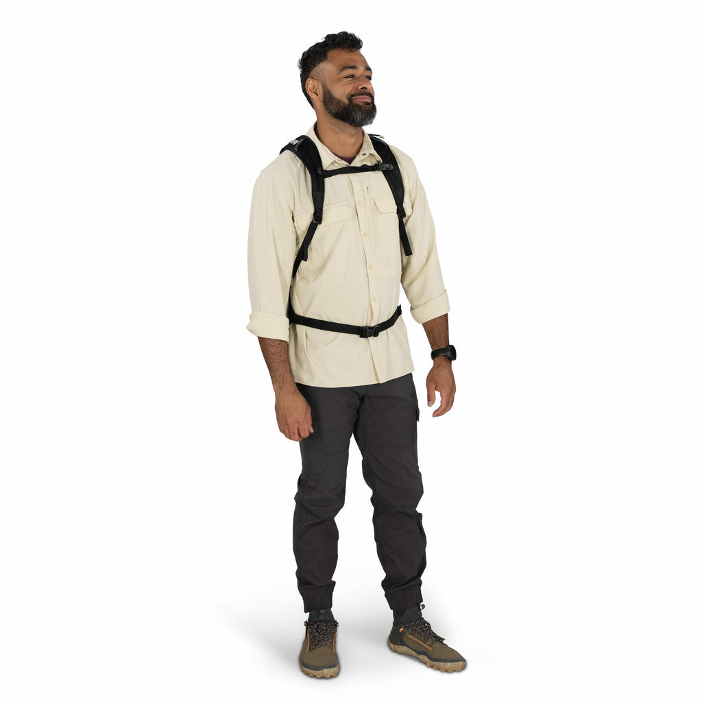 The image depicts a man dressed in outdoor attire suitable for hiking or trekking. He is wearing a light-colored long-sleeve shirt and dark cargo pants. On his feet, he has sturdy hiking shoes designed for rough terrain. A black backpack with visible stra