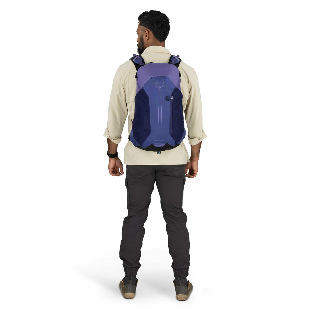 The image shows a man standing with his back to the camera, wearing outdoor clothing suitable for hiking or trekking. He is wearing a beige long-sleeve shirt with the sleeves rolled up and charcoal grey cargo pants, paired with sturdy brown hiking boots.