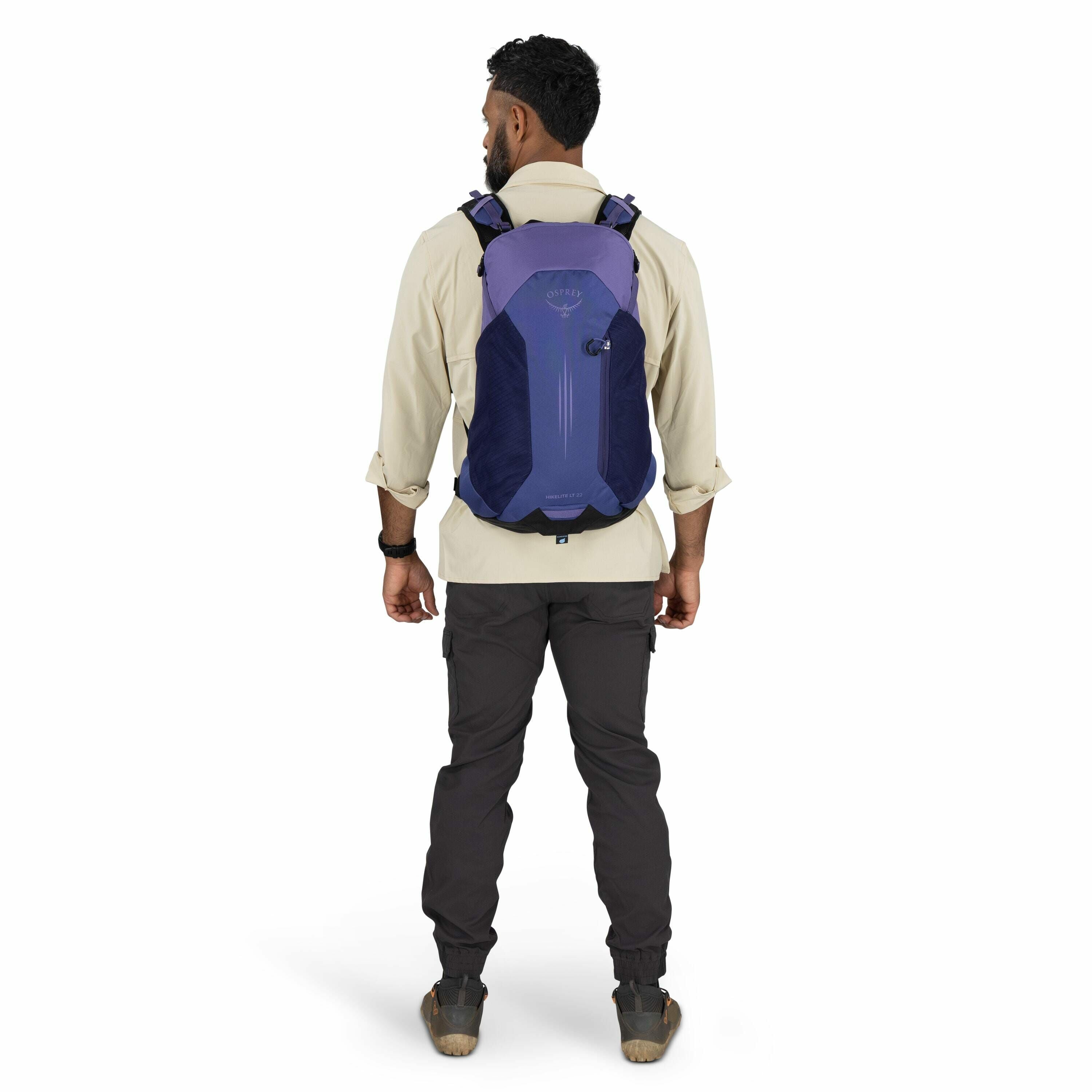 The image shows a man standing with his back to the camera, wearing outdoor clothing suitable for hiking or trekking. He is wearing a beige long-sleeve shirt with the sleeves rolled up and charcoal grey cargo pants, paired with sturdy brown hiking boots.