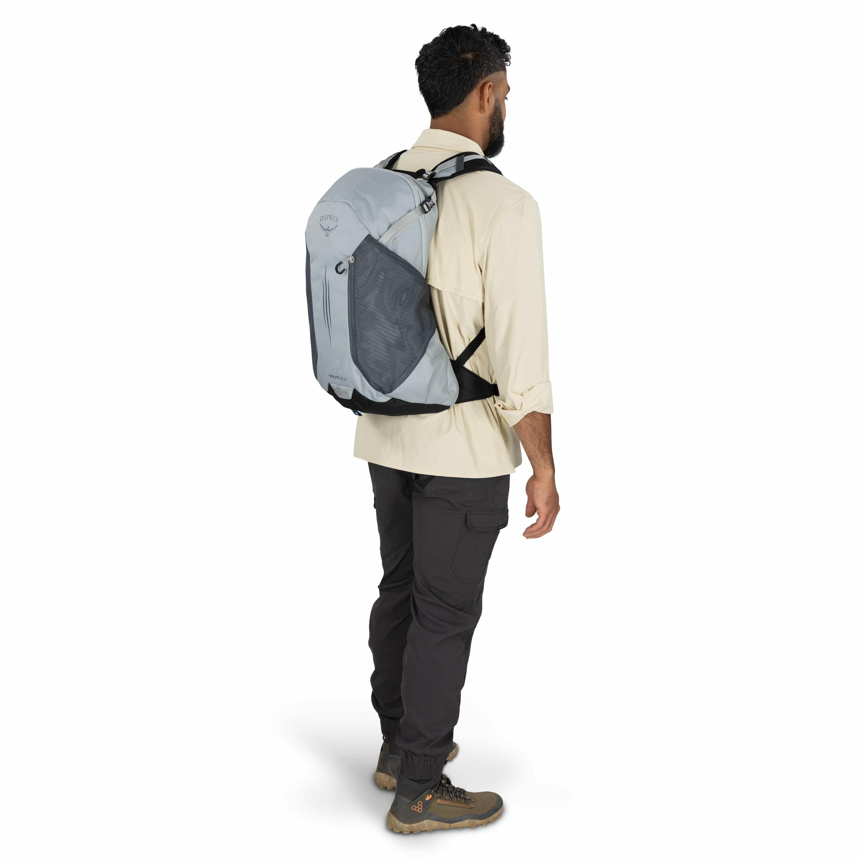 The image features a man standing and facing to the right, wearing a light gray backpack branded with the Osprey logo and the model name "Mira 22." He is dressed in a beige long-sleeve shirt with the sleeves rolled up and dark gray cargo pants, paired wit