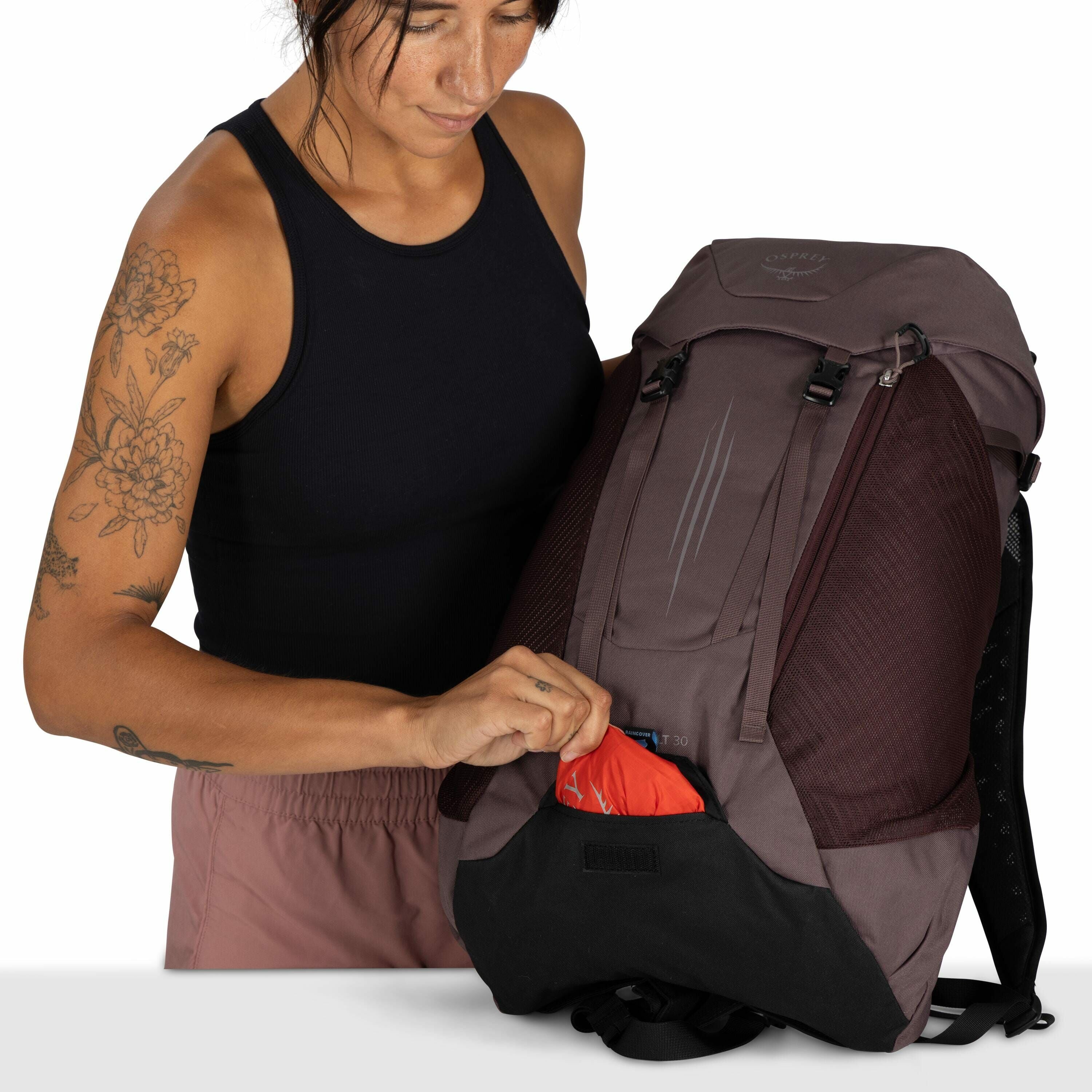 The image shows a person with tattoos wearing a sleeveless black top and light brown shorts, holding an Osprey backpack. The backpack is a dark brown color, equipped with mesh side panels and visible buckles at the top for securing the flap. The individua