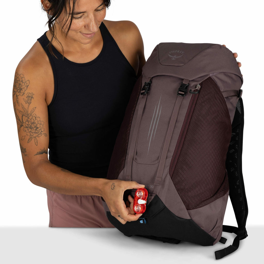 The image shows an individual holding a large backpack with the logo "Osprey" on the top flap. The backpack is mainly dark gray with maroon accents and features a streamlined design, with multiple compartments and straps. The person is holding a small red