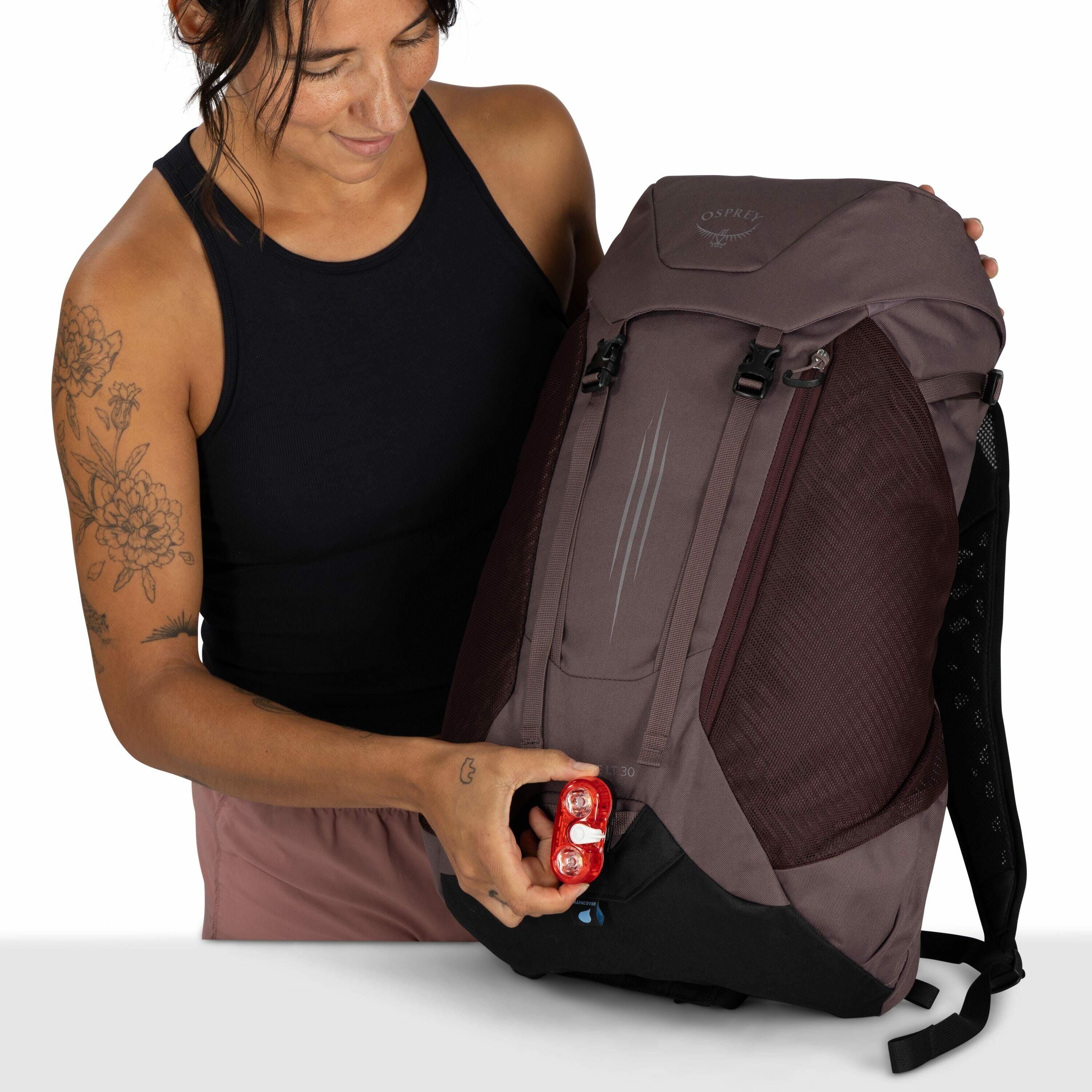 The image shows an individual holding a large backpack with the logo "Osprey" on the top flap. The backpack is mainly dark gray with maroon accents and features a streamlined design, with multiple compartments and straps. The person is holding a small red