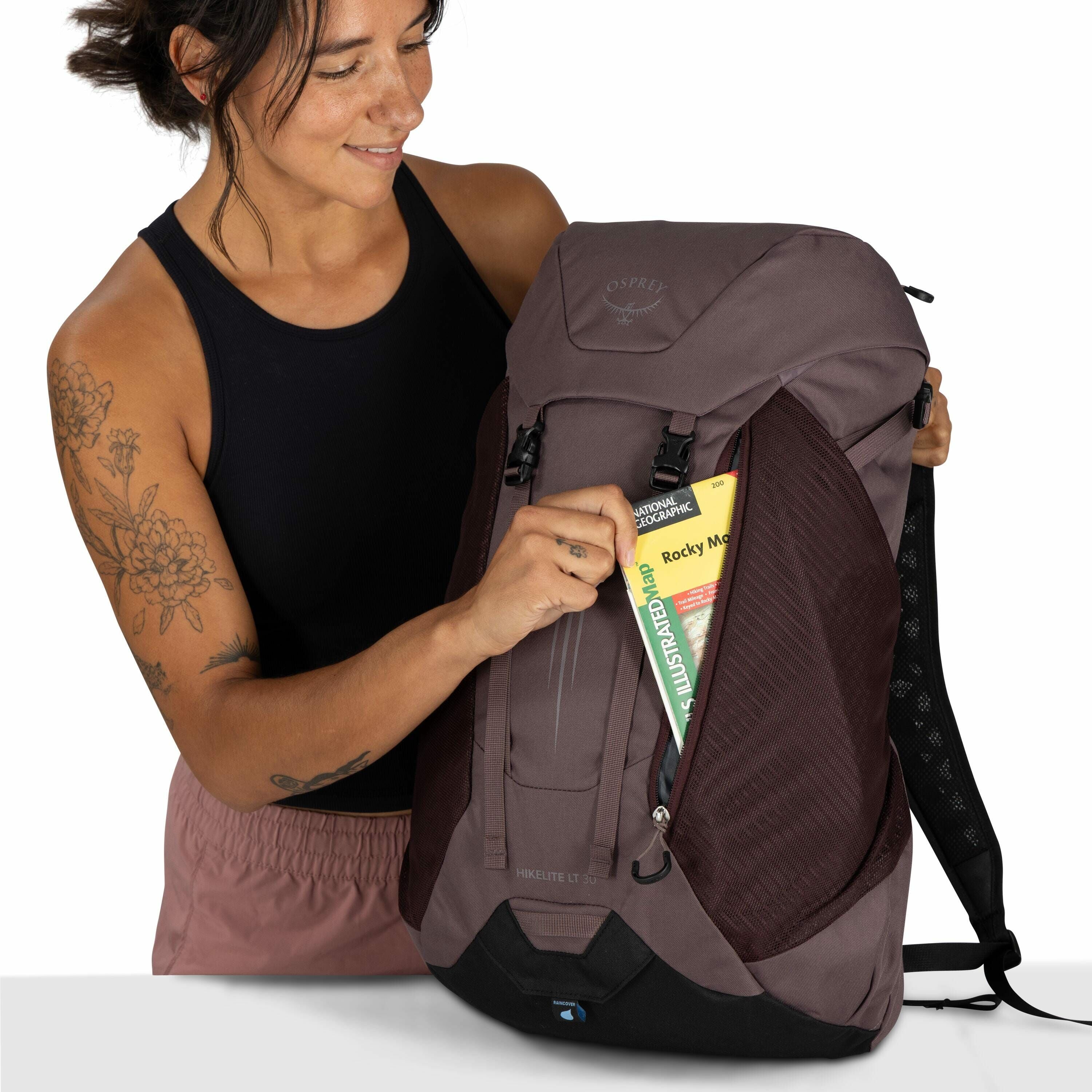 The image depicts a person holding a brown Osprey backpack in a studio setting. The person is dressed in a black tank top and mauve-colored shorts and has tattoos on both arms. The backpack features a front pocket being opened, revealing a National Geogra