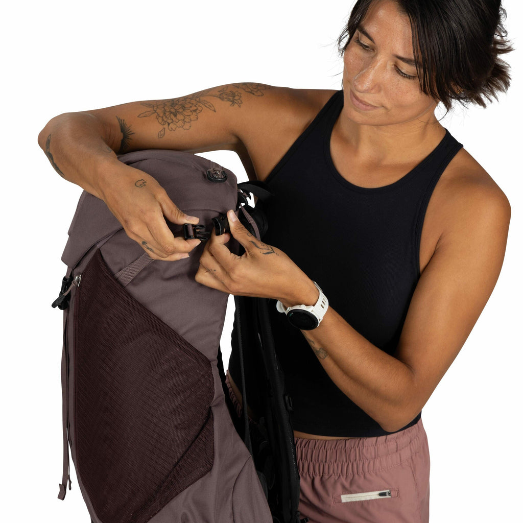 The image features a person adjusting the clasp on a backpack. The backpack is a muted burgundy color with a mesh pocket on its side, and the person is wearing a sleeveless black top and mauve shorts. The person has several tattoos on their arms, includin