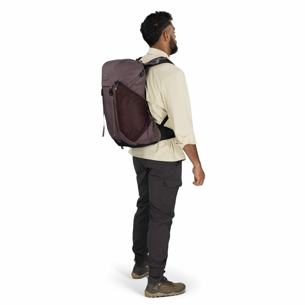 The image shows a man standing in a side profile view, wearing outdoor clothing and gear. He is dressed in a light beige long-sleeve shirt and dark gray cargo pants. He is also wearing hiking boots with olive green accents and gray socks. The man has a la