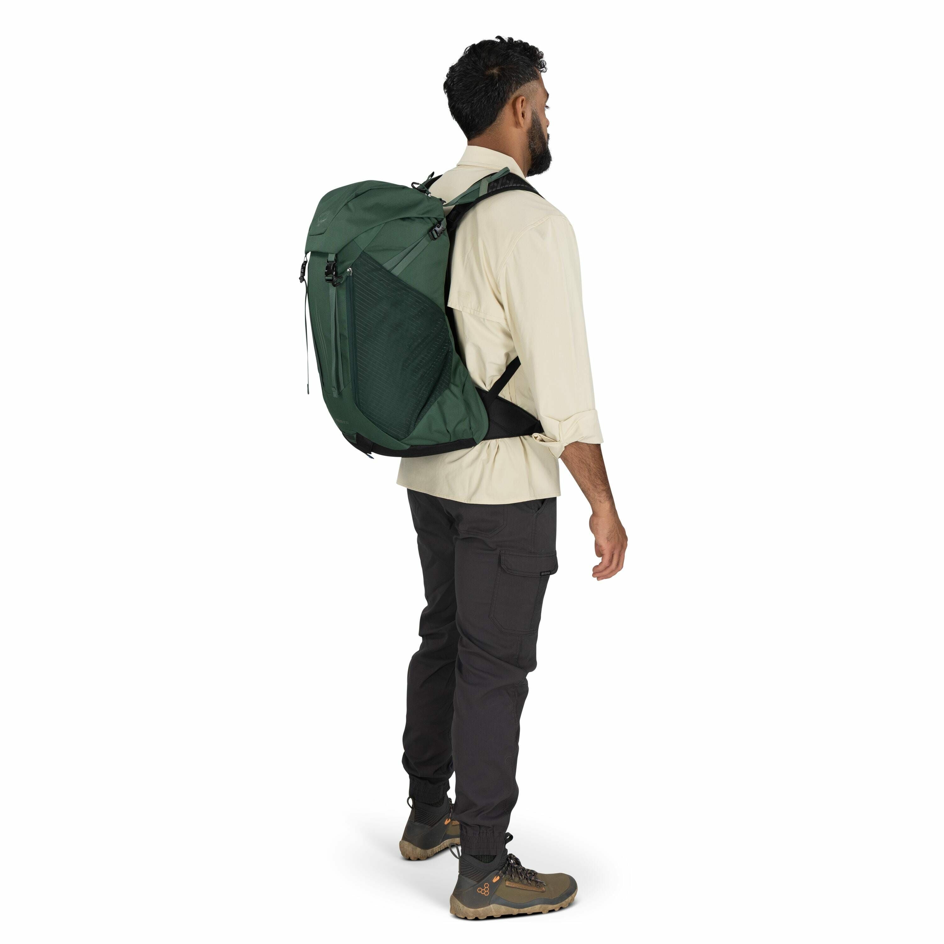 The image shows a man standing sideways, showcasing a green backpack. He is wearing an off-white long-sleeve shirt with rolled-up sleeves and dark grey cargo pants, complemented by olive green hiking boots. The backpack features multiple compartments and