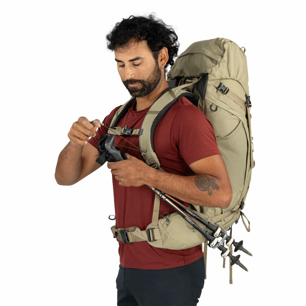 The image shows a person wearing a large, light brown backpack equipped with multiple straps and pockets. They are holding trekking poles, which are attached to the side of the backpack. The individual is dressed in a red short-sleeve shirt and dark pants
