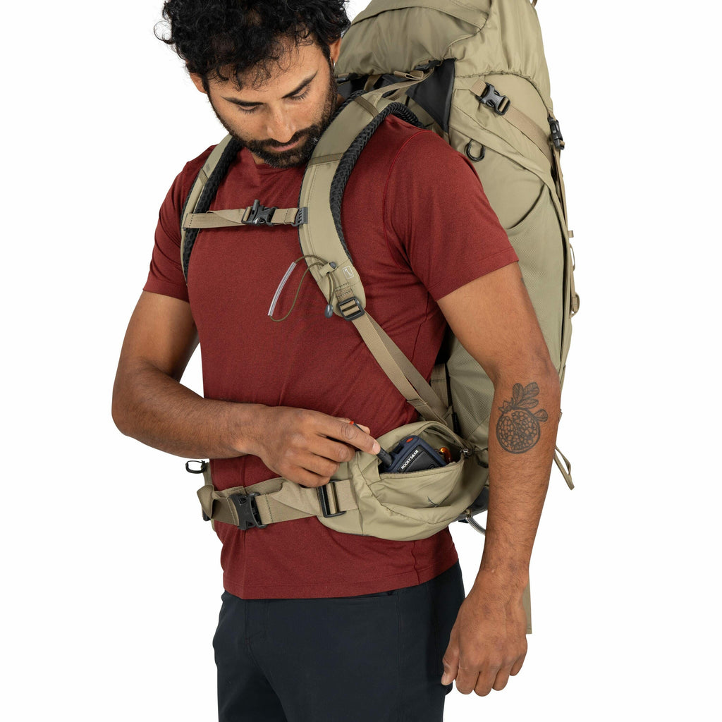 The image shows a man wearing a large, tan-colored backpack designed for outdoor activities, such as hiking or camping. He is dressed in a red t-shirt and dark pants, accessorized with a tattoo of a fruit resembling a pomegranate on his left forearm. The