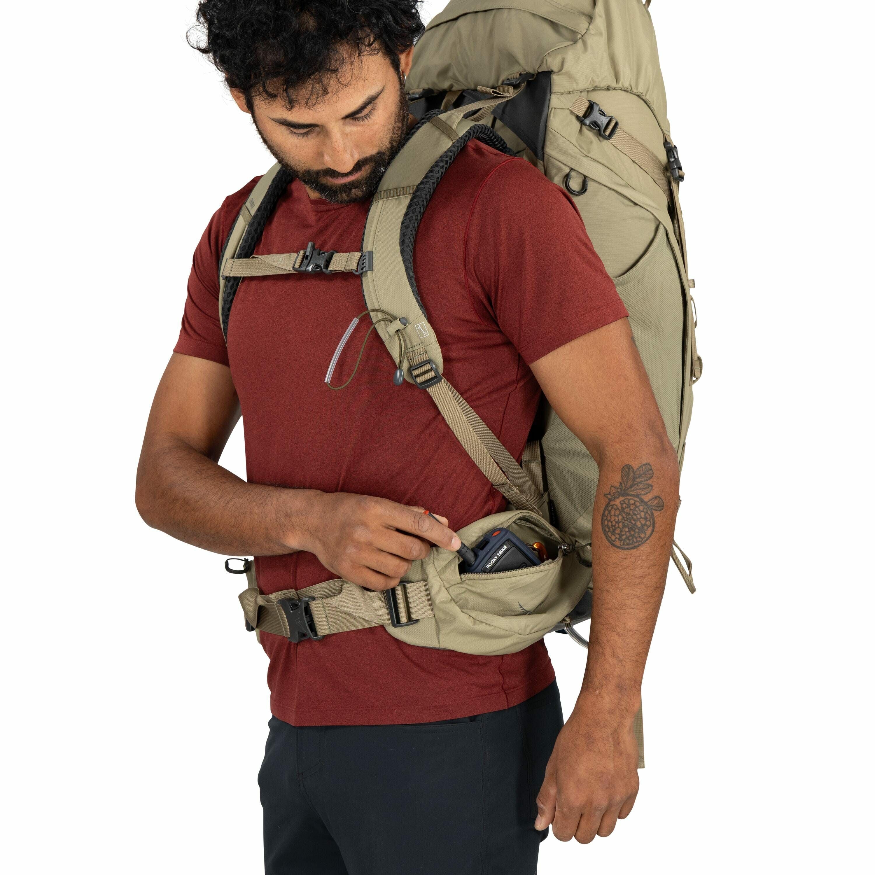 The image shows a man wearing a large, tan-colored backpack designed for outdoor activities, such as hiking or camping. He is dressed in a red t-shirt and dark pants, accessorized with a tattoo of a fruit resembling a pomegranate on his left forearm. The