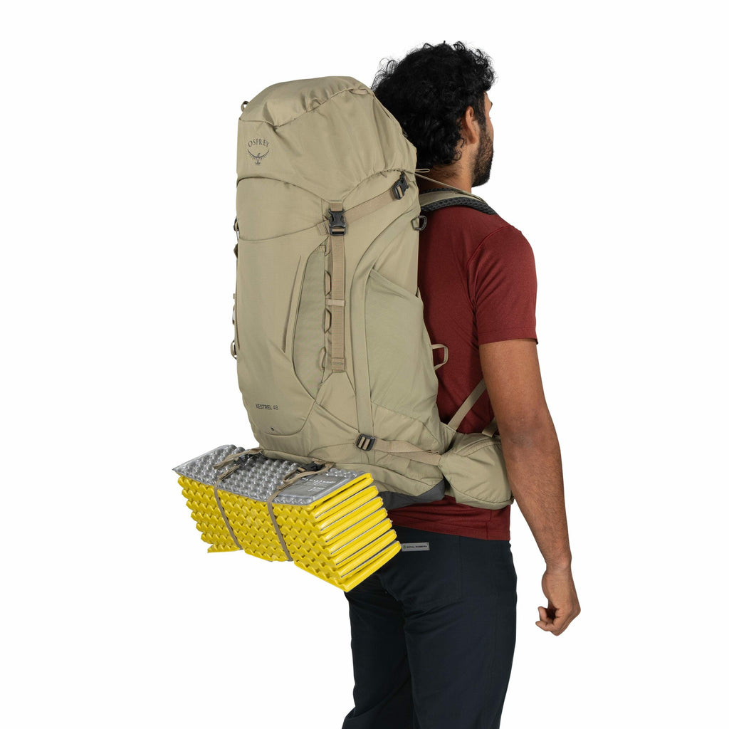 The image shows a man wearing a large tan hiking backpack by Osprey, with the model name "Kestrel 48" printed on the side. He has a rolled-up yellow and silver foam sleeping pad strapped horizontally to the bottom of the backpack. The man is dressed in a