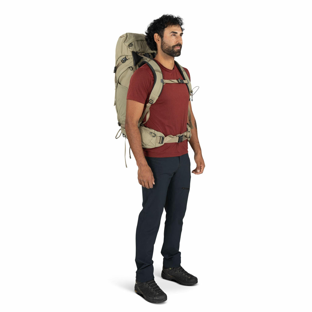 The image shows a person standing upright, wearing a casual outdoor outfit suitable for hiking or trekking. They are dressed in a red short-sleeve t-shirt, dark-colored outdoor pants, and black hiking shoes. The person has a large beige backpack secured w