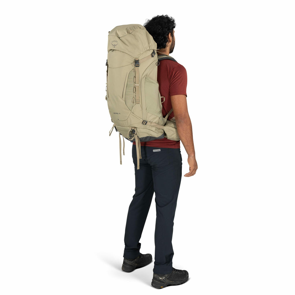 The image shows a man with dark, curly hair wearing an Osprey hiking backpack. The backpack is beige and appears to be a large-capacity model, designed for extended trips, evident by its tall and sturdy structure. The man is dressed in a short-sleeved mar