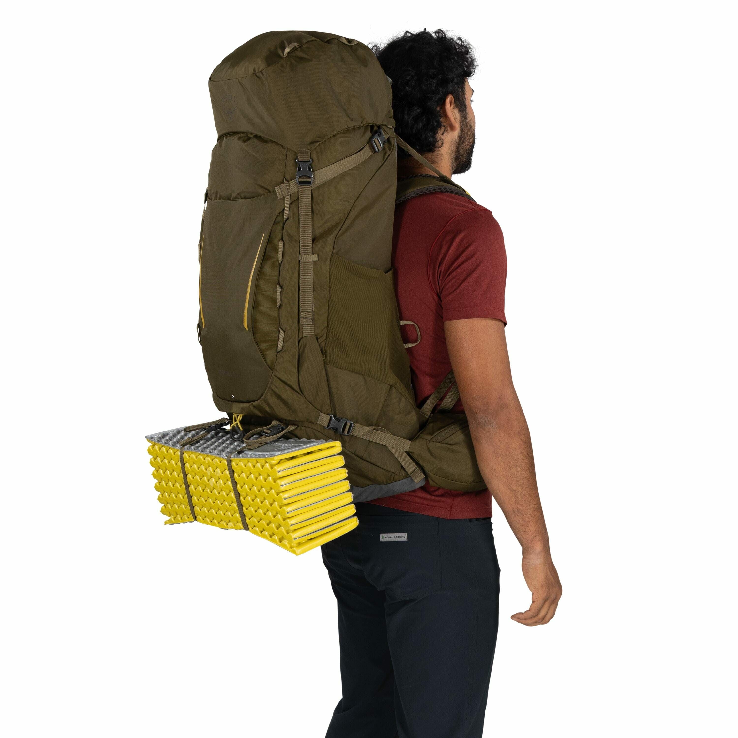 The image shows a person with dark curly hair, dressed in a red short-sleeved shirt and dark pants, carrying a large green backpack. Attached to the bottom of the backpack is a folded yellow and silver sleeping pad, secured with straps. The backpack appea