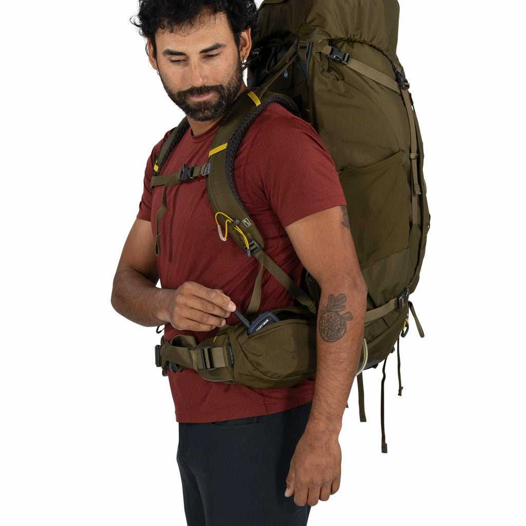 The image depicts a person wearing a burgundy short-sleeve shirt and black pants, equipped with a large olive-green backpack. The backpack is strapped on securely with shoulder and waist straps, indicating it is intended for heavy loads or extended hikes.