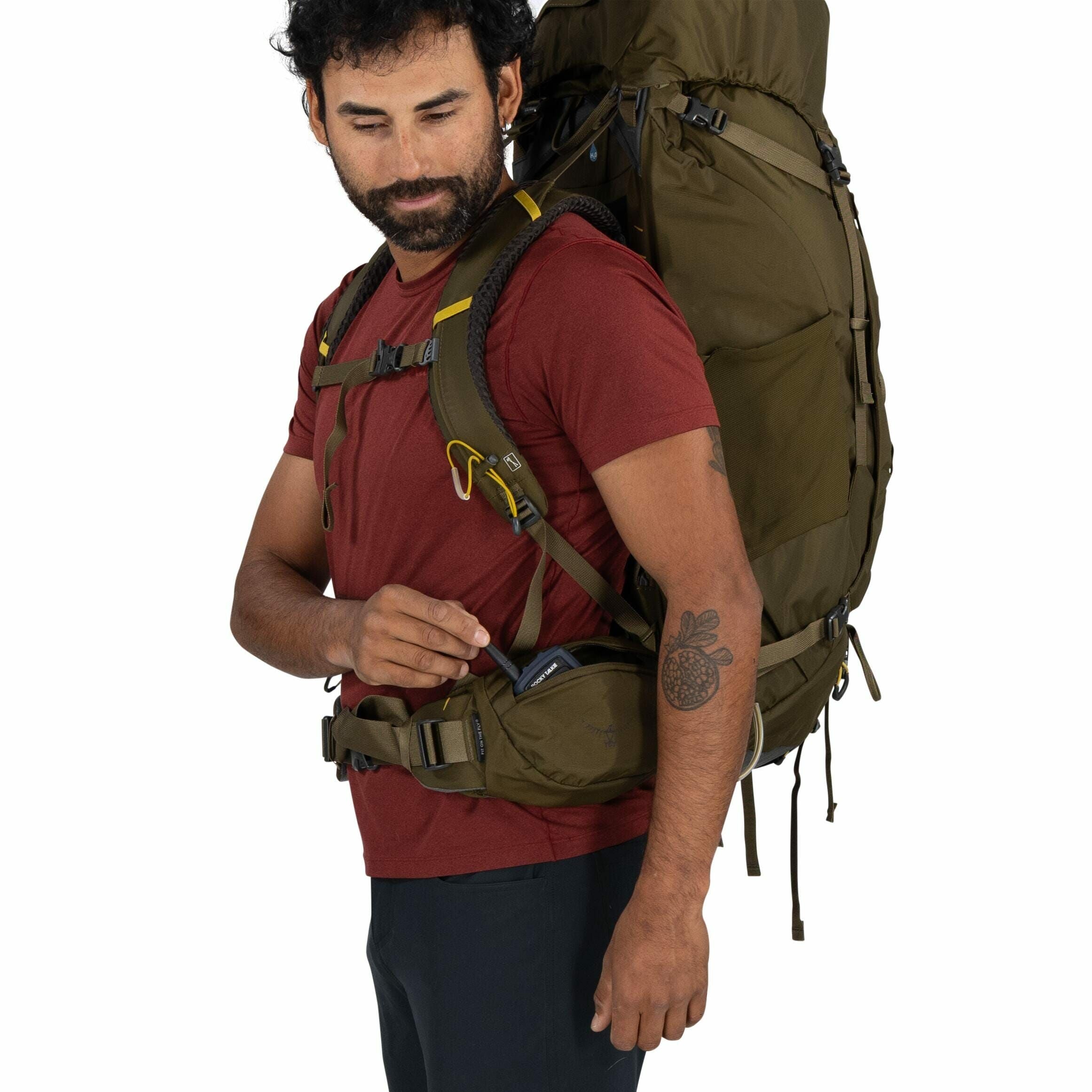 The image depicts a person wearing a burgundy short-sleeve shirt and black pants, equipped with a large olive-green backpack. The backpack is strapped on securely with shoulder and waist straps, indicating it is intended for heavy loads or extended hikes.