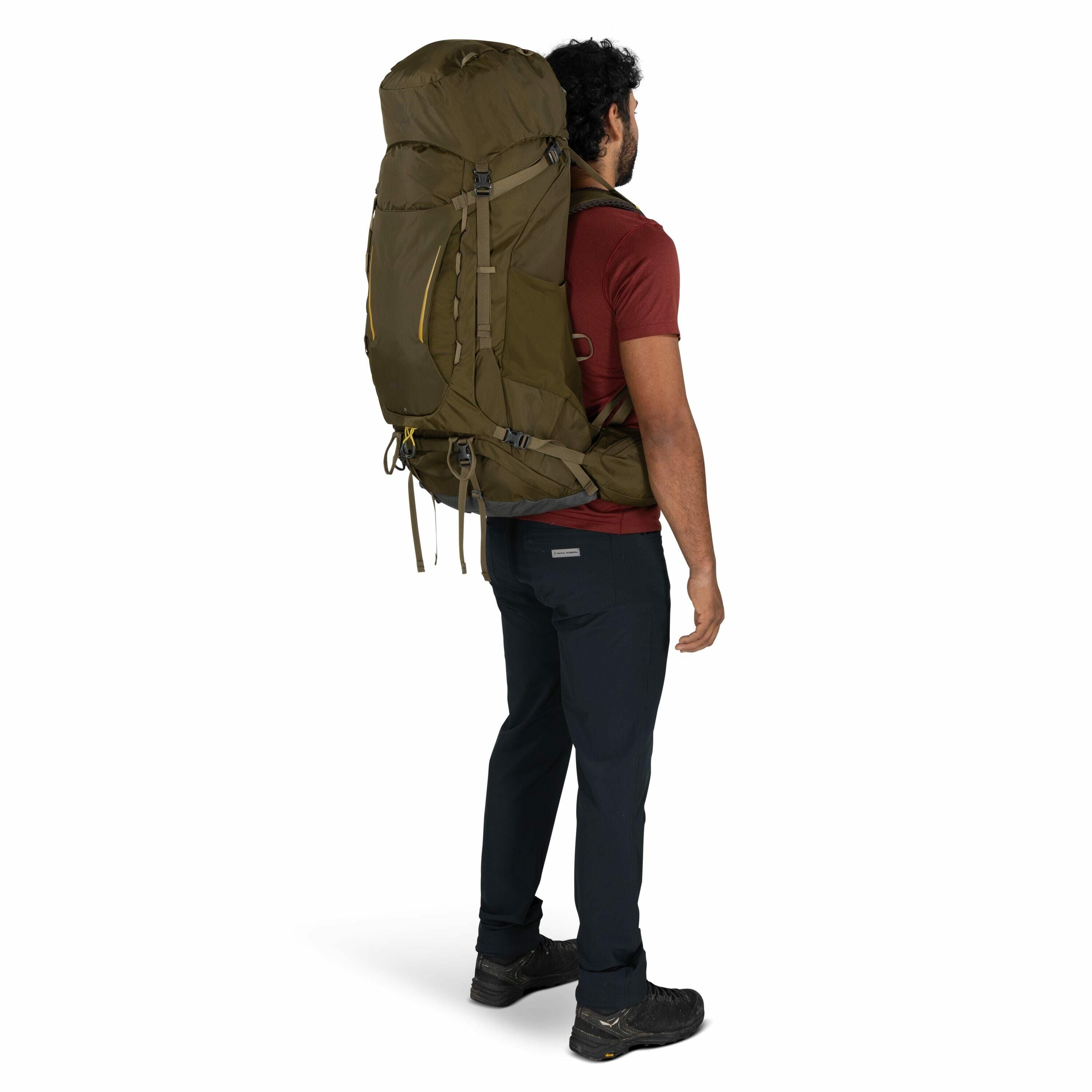 The image shows a man standing and facing sideways, carrying a large, olive-green hiking backpack. He is dressed in a maroon t-shirt and dark navy pants, accompanied by sturdy, black hiking shoes, ready for outdoor adventure. The backpack is notably large