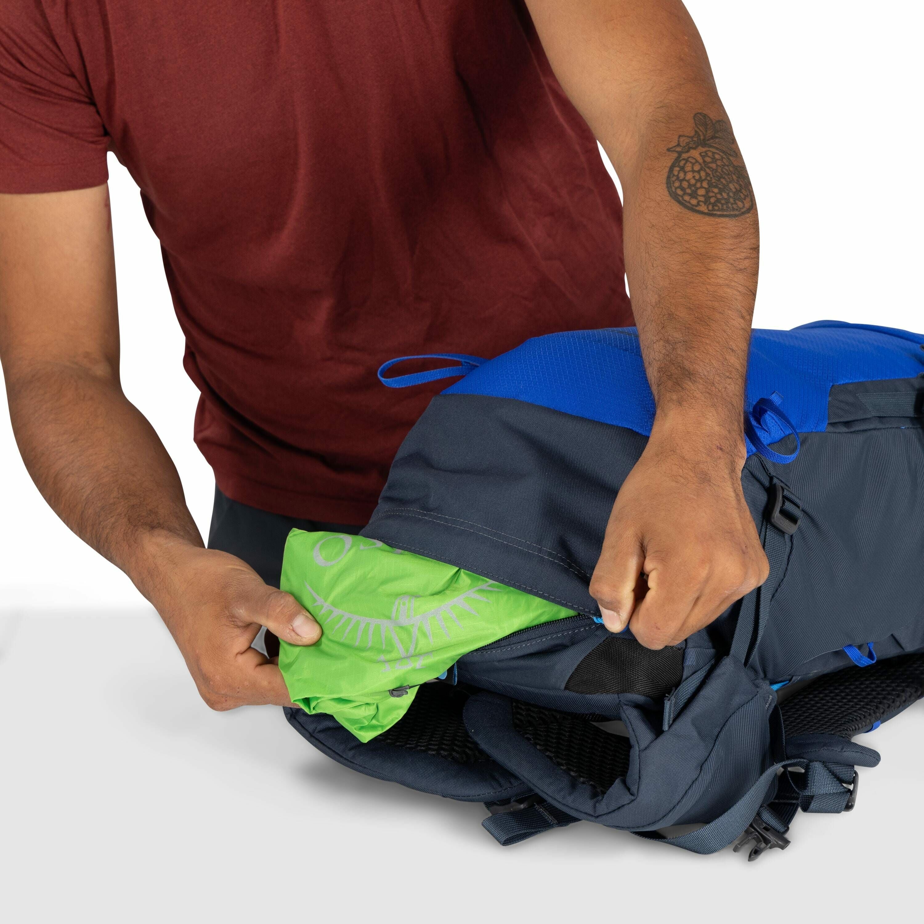 The image depicts a person wearing a red shirt demonstrating how to insert or remove a green fabric with a logo from a side pocket of a blue and black backpack. The personÕs left arm is visible, showing a tattoo of a pomegranate. The green fabric appears