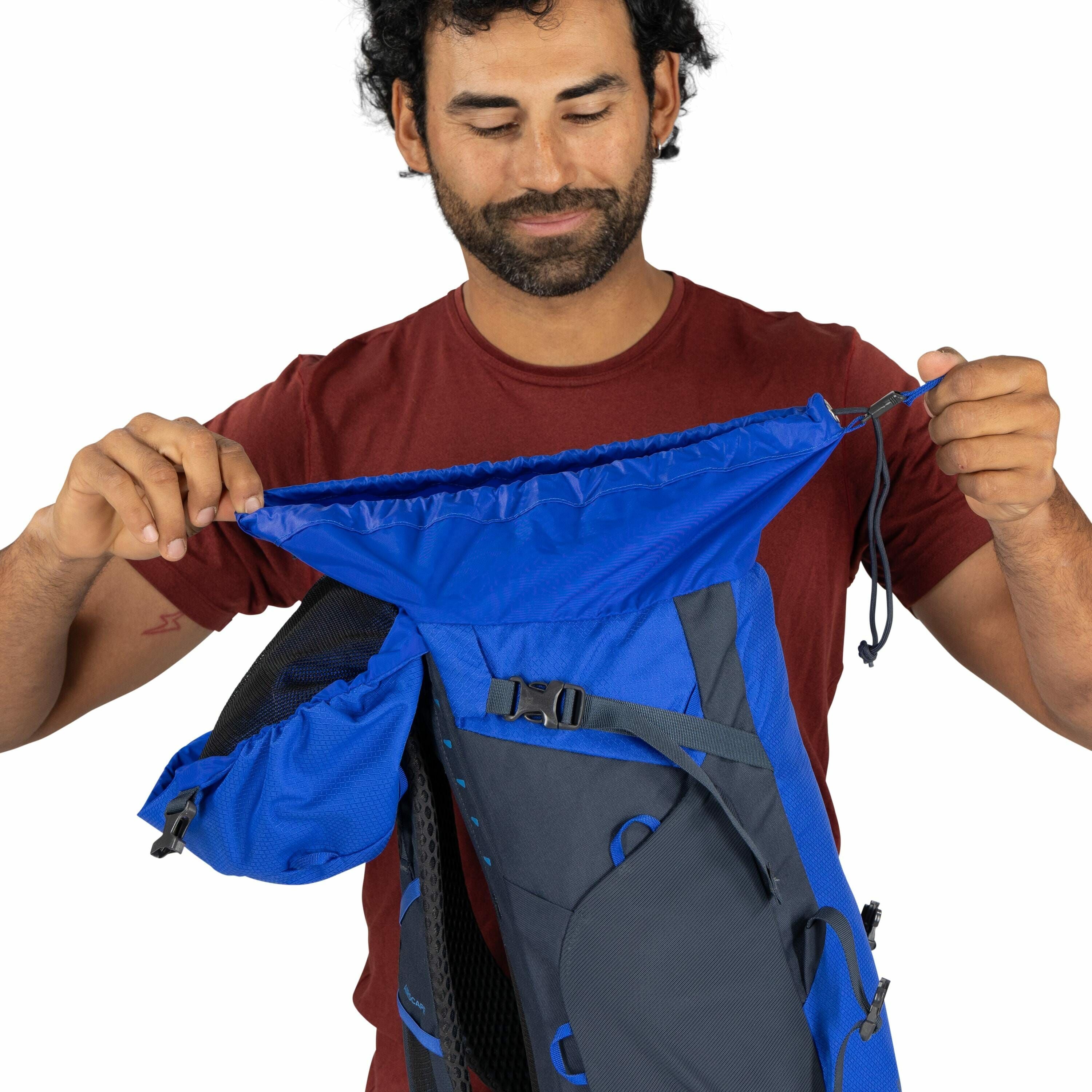 The image shows a person wearing a maroon t-shirt, holding a blue and gray backpack. The backpack appears to have a large main compartment with a drawstring closure, which the person is opening. There are several details such as buckles and straps visible