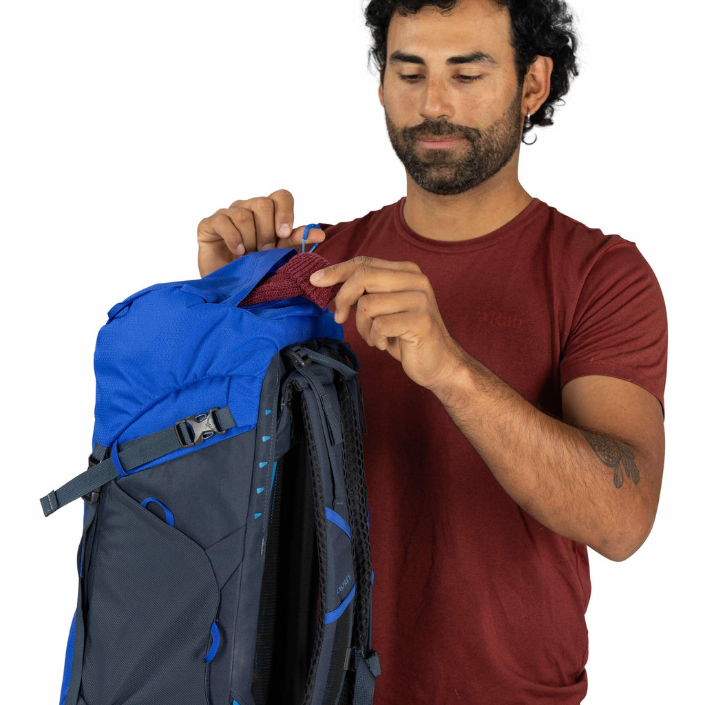 The image depicts a person wearing a maroon t-shirt with a subtle "Rab" logo on the chest. They are interacting with a blue and dark grey backpack, likely an outdoor or hiking pack, as indicated by its design and features. The person is holding a small, m