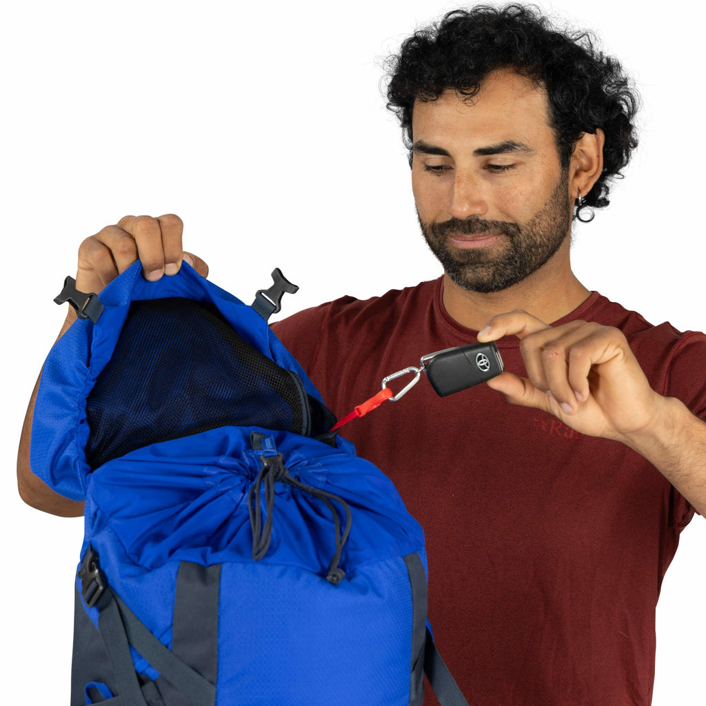 The image shows a person demonstrating a feature of a blue backpack. The backpack is open, and the person is holding a black car key fob, identifiable by the Toyota logo, which is attached to a red carabiner clip. This clip is fastened to a loop inside th