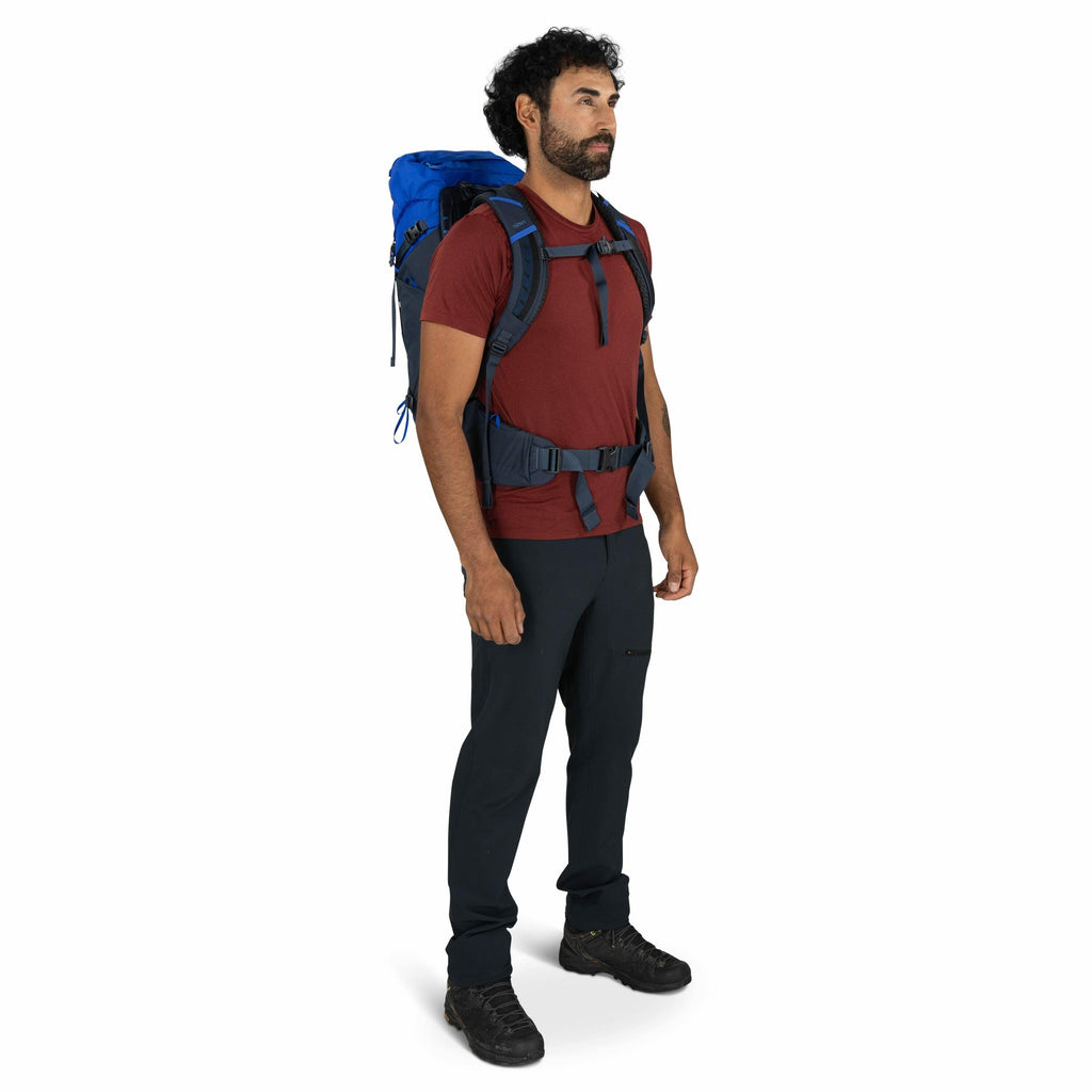 The image shows a person dressed in hiking gear, standing in a three-quarter view. They are wearing a maroon t-shirt paired with dark hiking pants and black hiking shoes. The person is carrying a large blue backpack with padded straps, secured with both a