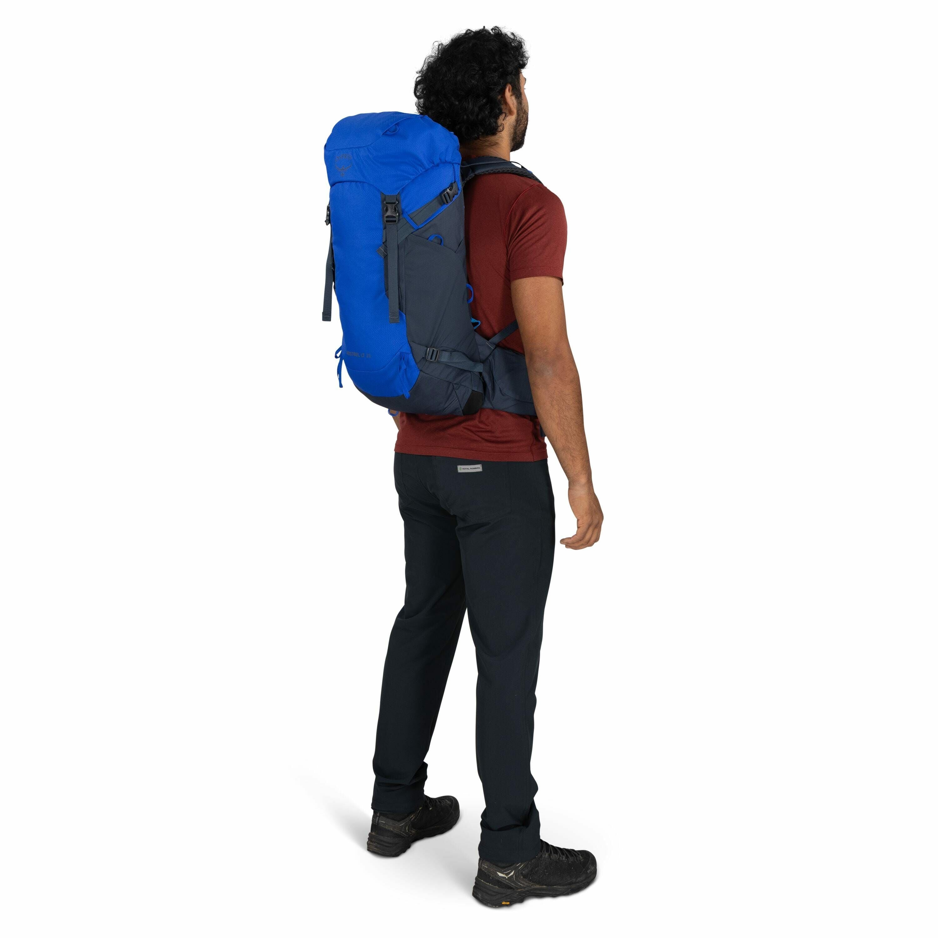 The image features a man standing with his back turned, wearing a large blue and dark gray backpack. He is dressed in outdoor attire, including a maroon t-shirt, black hiking pants, and sturdy hiking shoes. His curly hair is visible from the back, and he