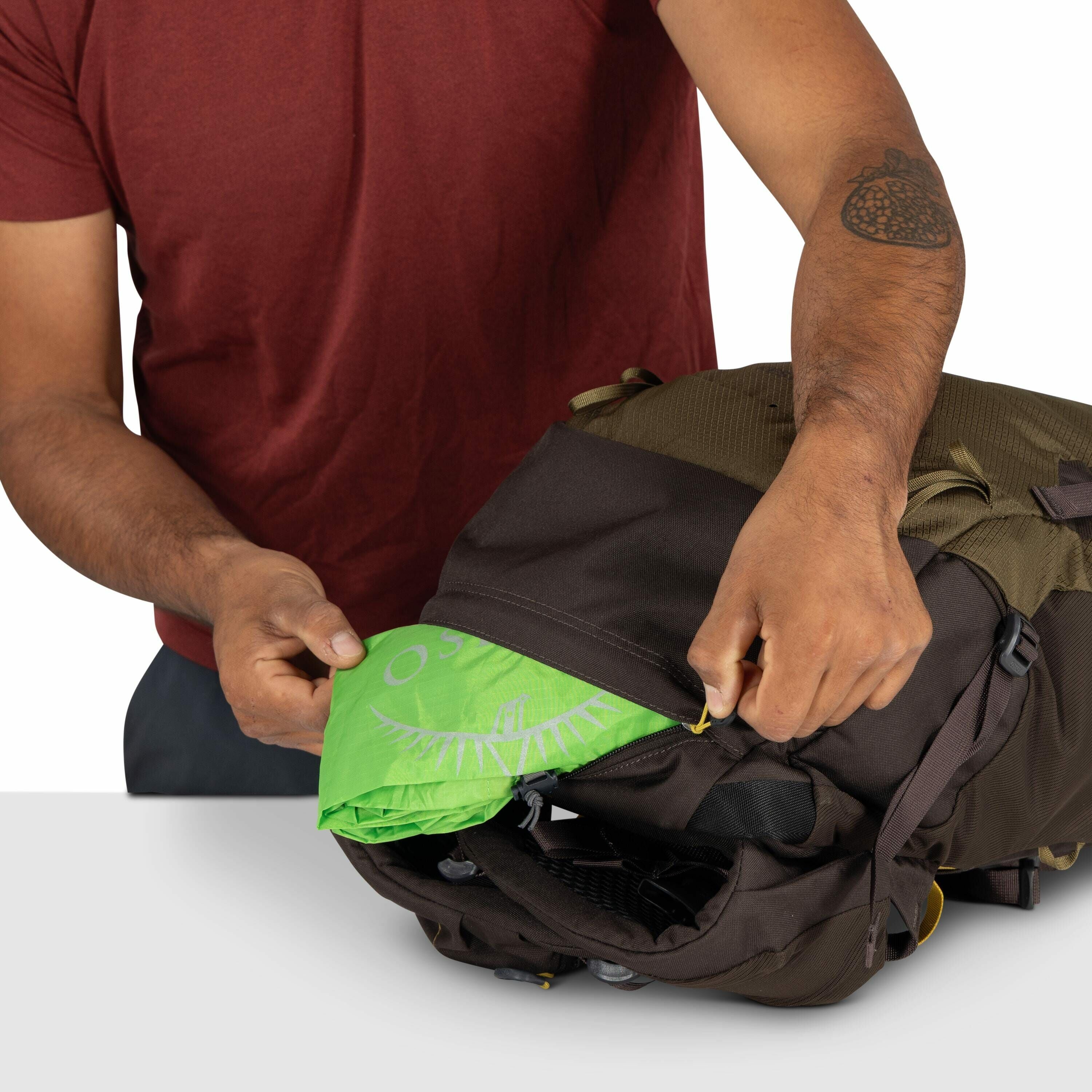 The image shows a person with medium skin tone wearing a red shirt, unpacking or packing a green fabric from a dark brown backpack. The fabric has a logo with the word "OSPREY" and a white design resembling a bird's wingspan, suggesting it is likely a rai