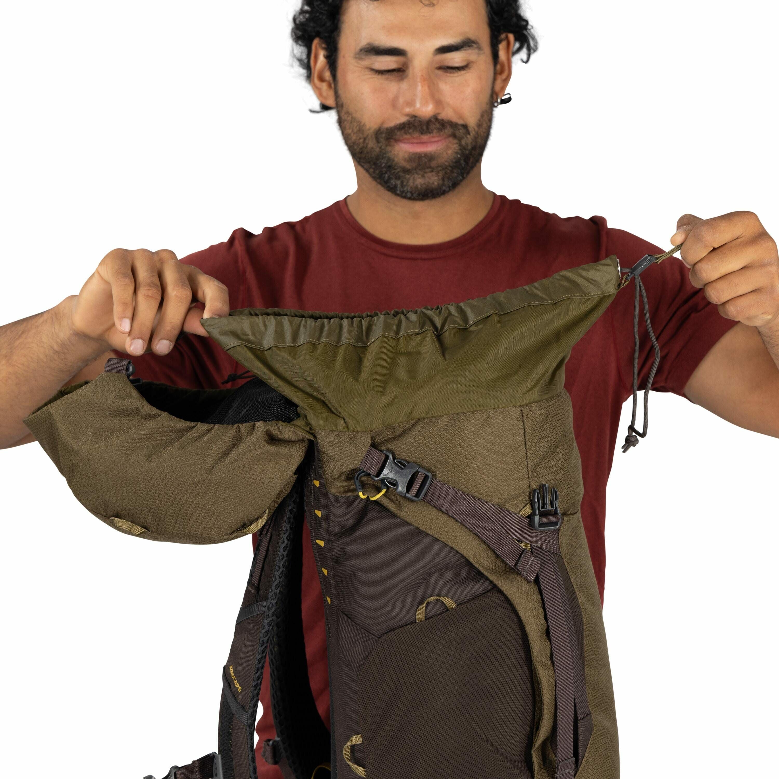 The image depicts a person wearing a red short-sleeved shirt, showcasing a brown and green backpack. The backpack features multiple compartments, including a spacious main compartment with an adjustable drawstring closure that the person is pulling to dem
