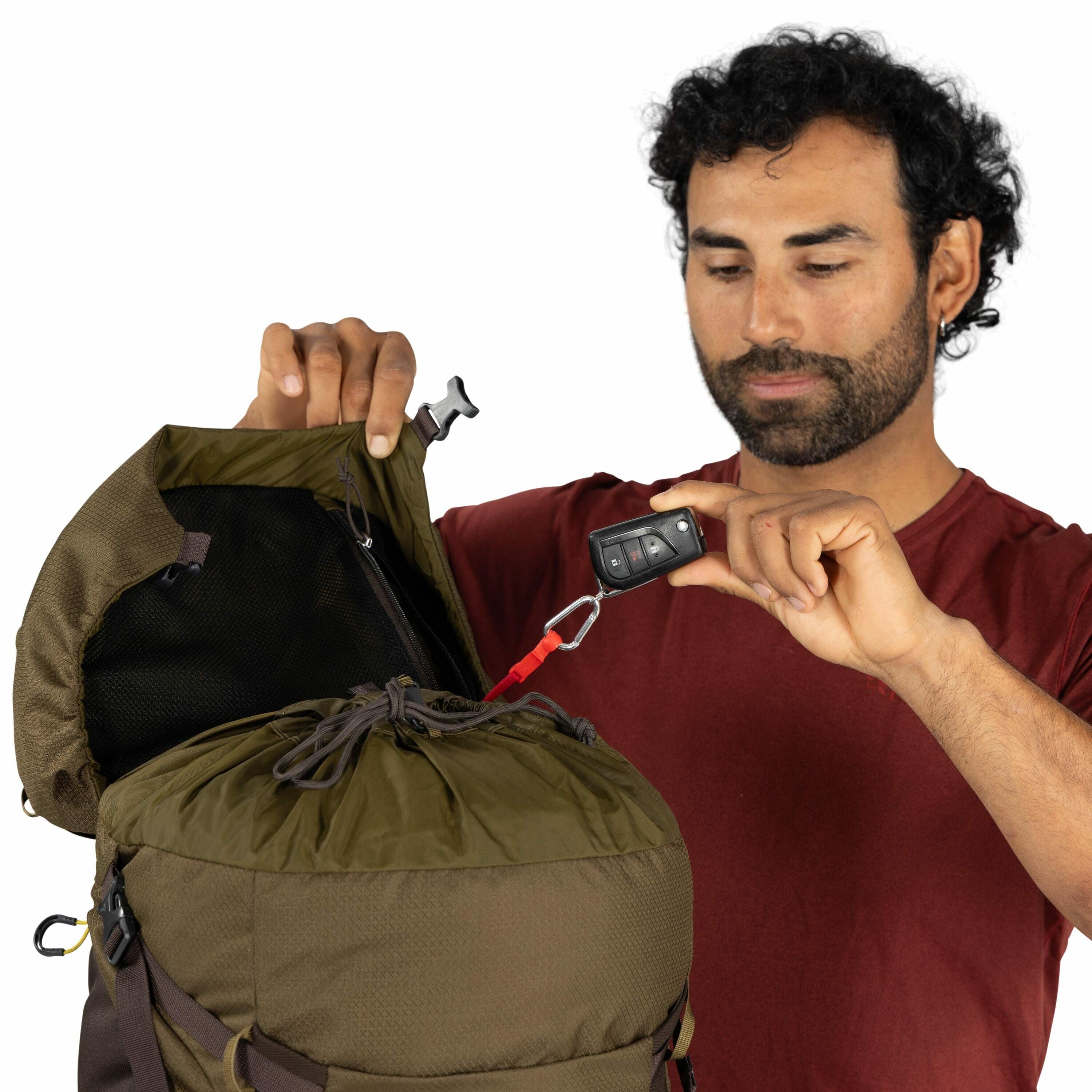 The image shows a person in a red shirt holding an olive-green backpack, which has its top compartment open. In one hand, the person is holding a car key fob attached to a small red strap and a metal carabiner clip. The interior of the backpack's top flap