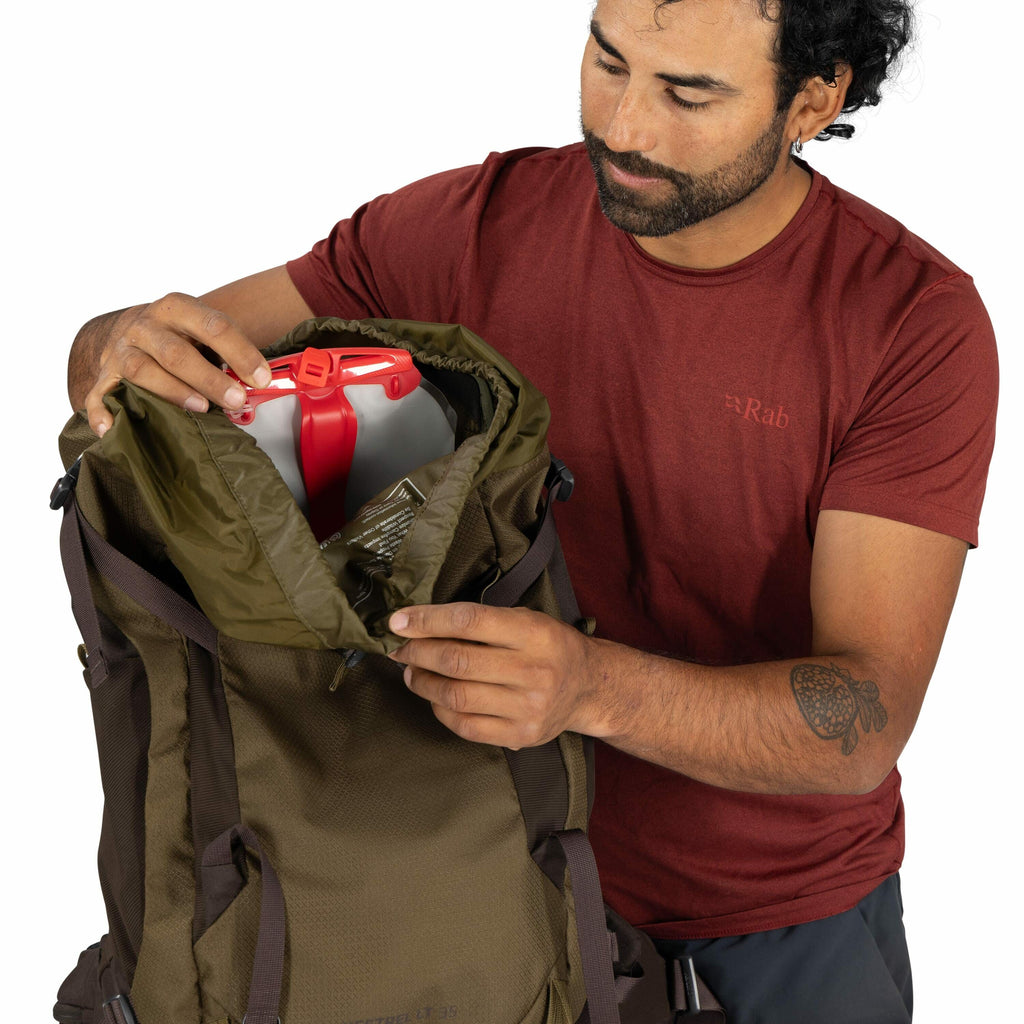 The image shows a person in a red Rab T-shirt, packing or unpacking a green and brown backpack. Inside the top section of the backpack, a red and white water container is visible. The person is using both hands to open the top of the backpack, which appea