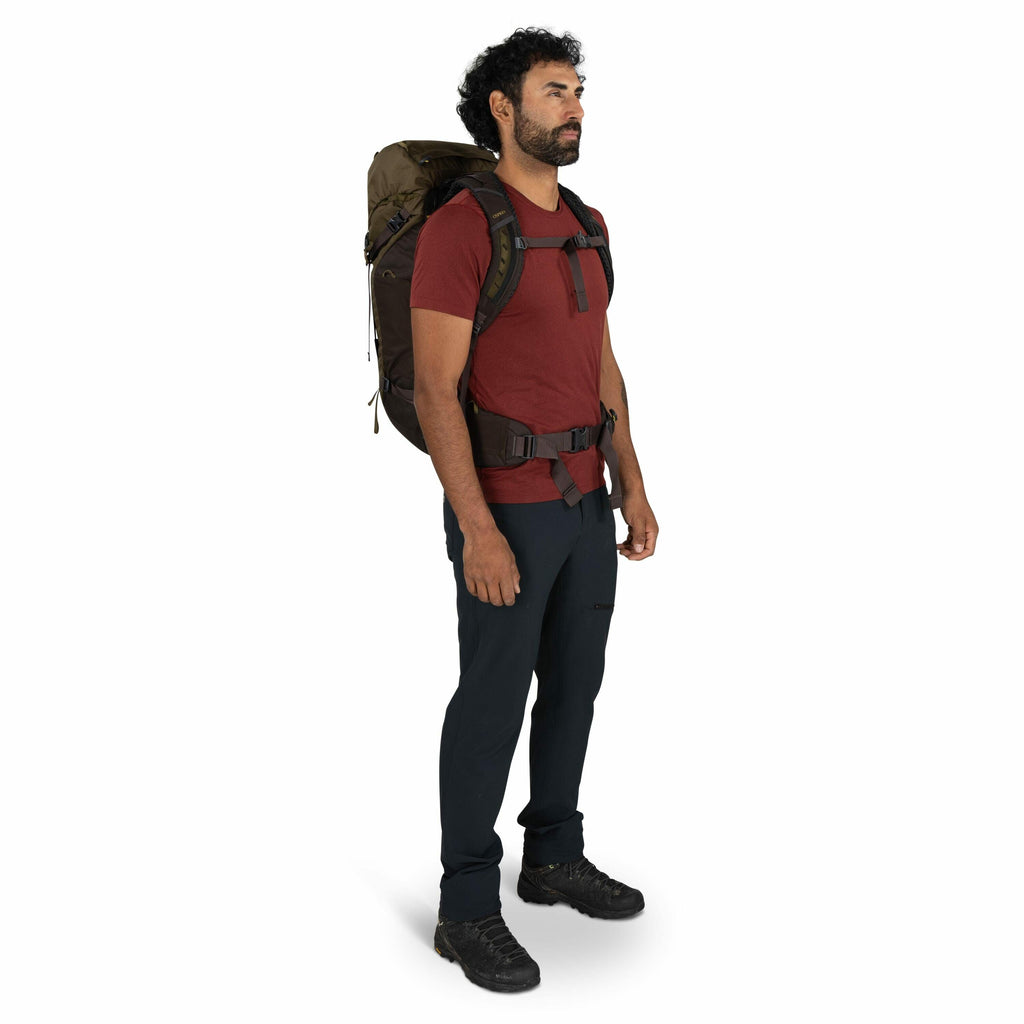 The image shows a person standing in a side profile view wearing outdoor hiking gear. The individual is dressed in a red short-sleeve T-shirt and dark navy hiking pants, paired with black outdoor shoes. They are carrying a large olive green hiking backpac