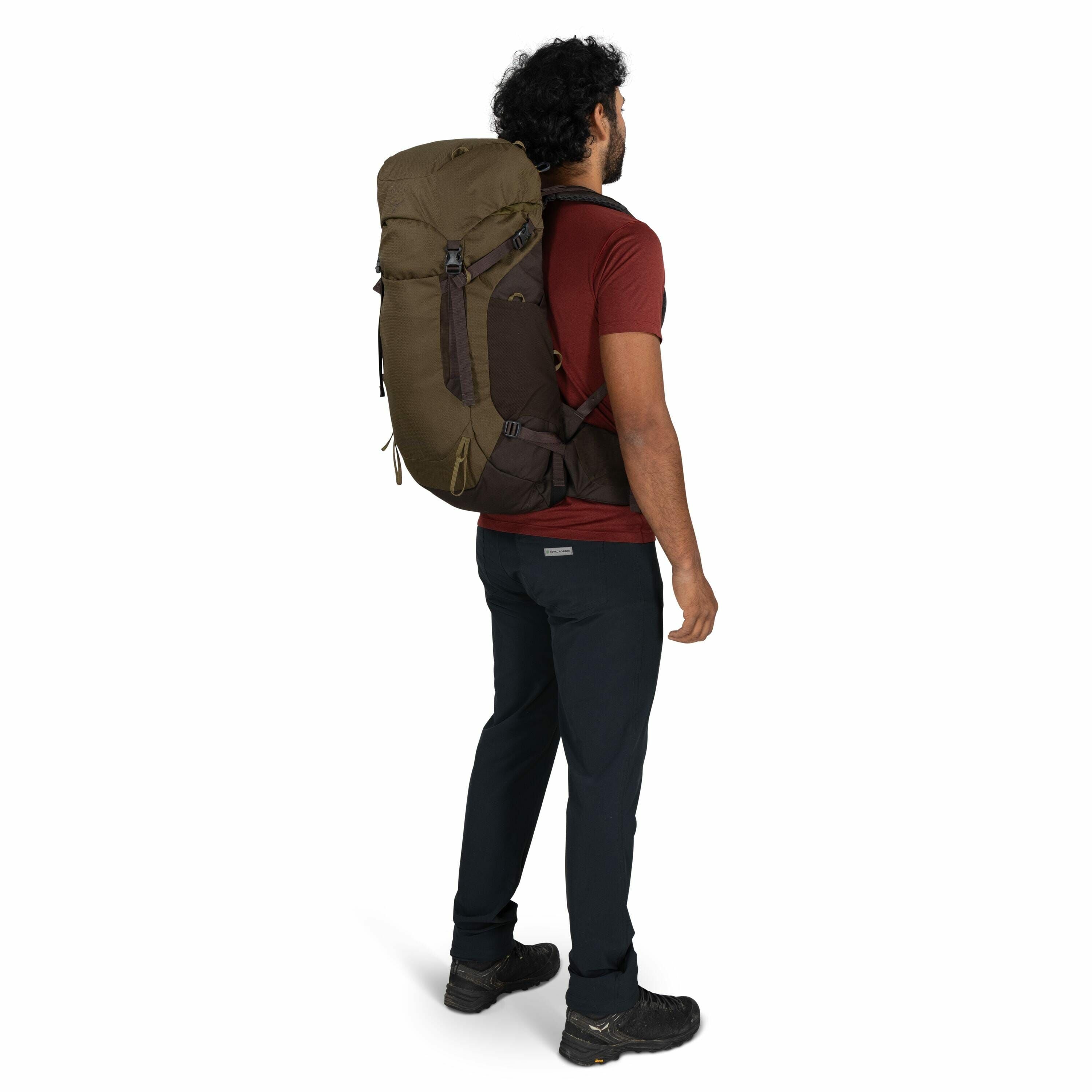 The image shows a person standing, viewed from the side, wearing a large olive-green and brown hiking backpack. The individual is dressed in a short-sleeve maroon t-shirt, black hiking pants, and black hiking shoes. They have short, curly black hair and a