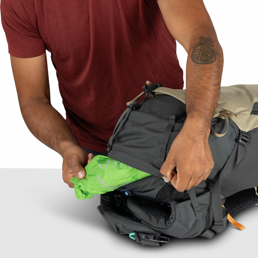The image shows a person wearing a maroon shirt and holding a backpack while pulling out a green item from one of its compartments. The green item seems to be a fabric cover with "Osprey" written on it, likely a rain cover for the backpack. The backpack i