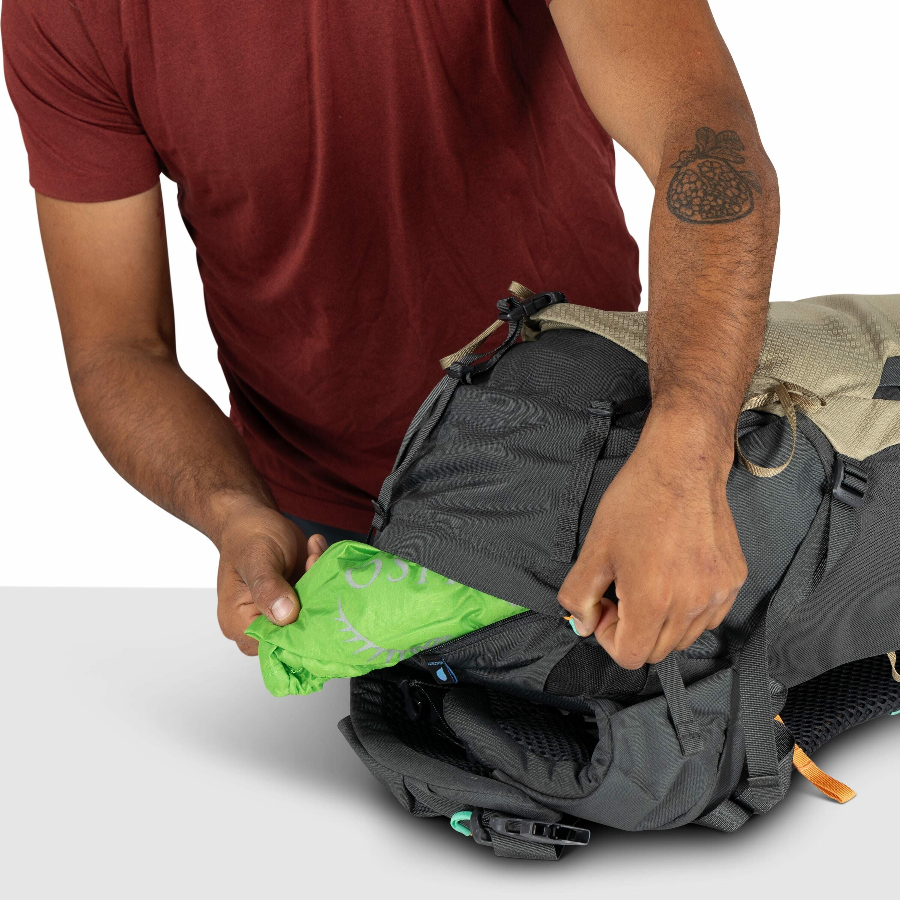The image shows a person wearing a maroon shirt and holding a backpack while pulling out a green item from one of its compartments. The green item seems to be a fabric cover with "Osprey" written on it, likely a rain cover for the backpack. The backpack i