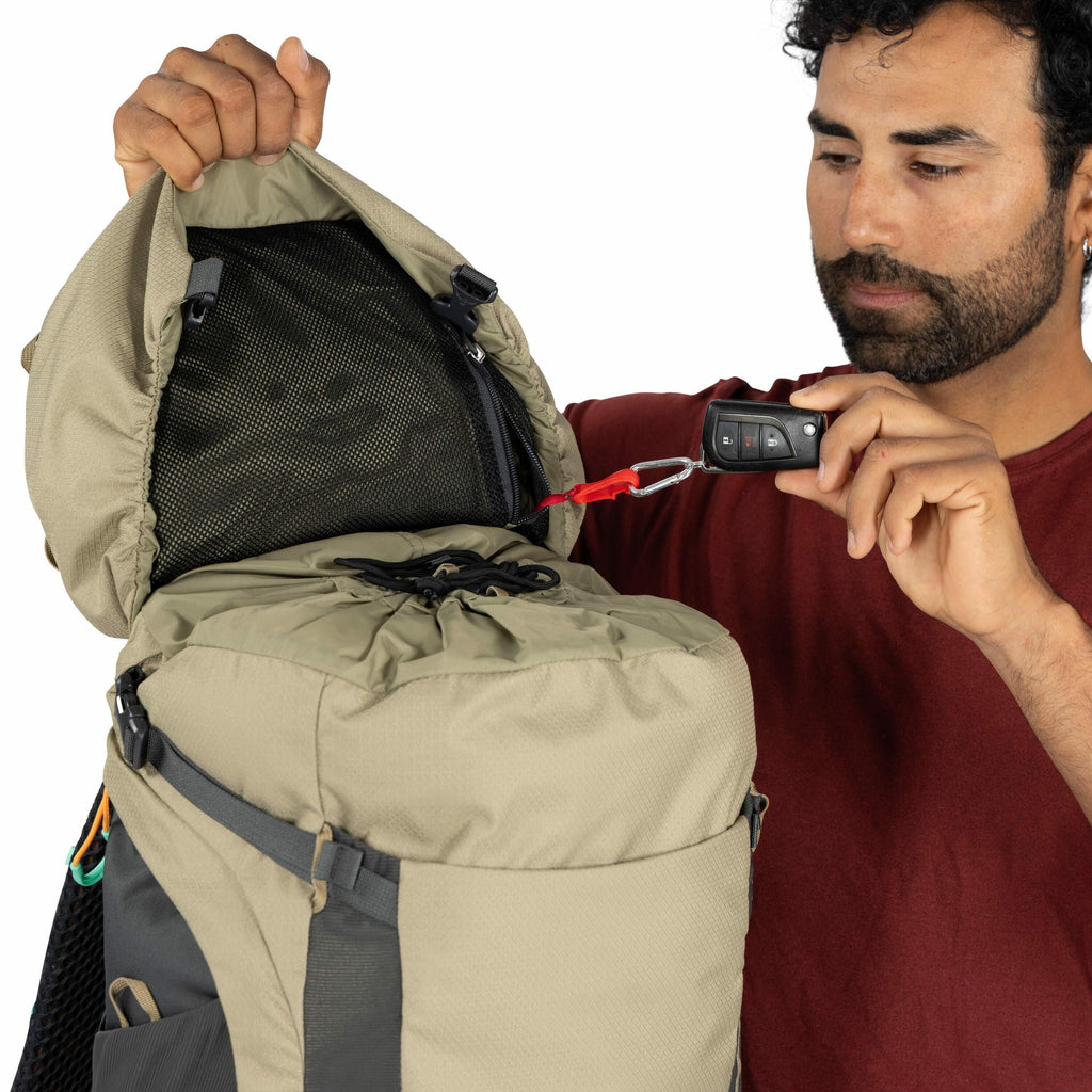The image shows a person holding a car key fob that is attached to a carabiner clip with a red strap, which is connected to the inside of a backpack. The backpack is open, revealing a black mesh pocket in the interior lid. The person is wearing a maroon s