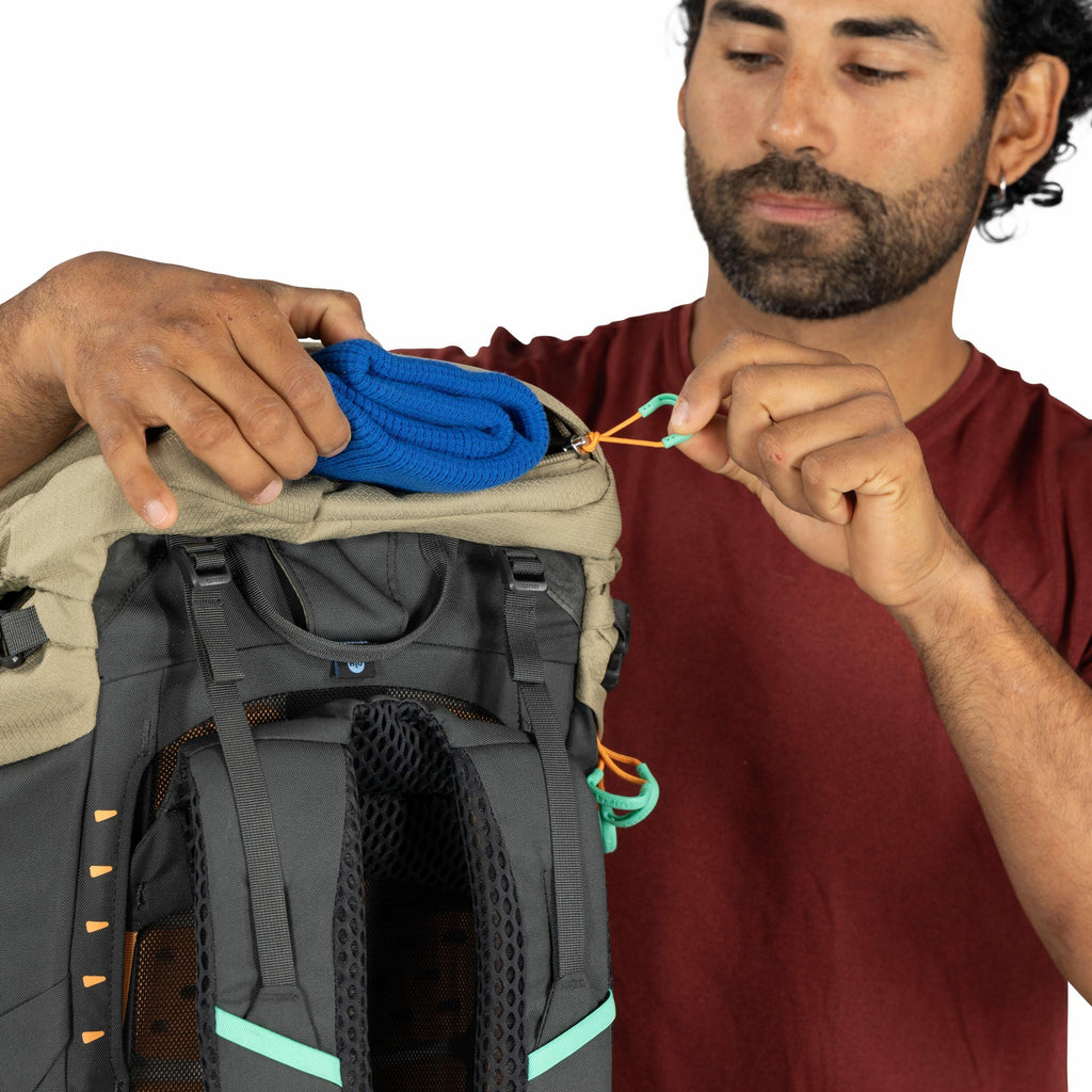 The image depicts a person adjusting the contents of a backpack, which has a color scheme of beige and black. The person is placing a rolled-up blue item, possibly a towel or blanket, into the top section of the backpack. They are seen pulling on an orang
