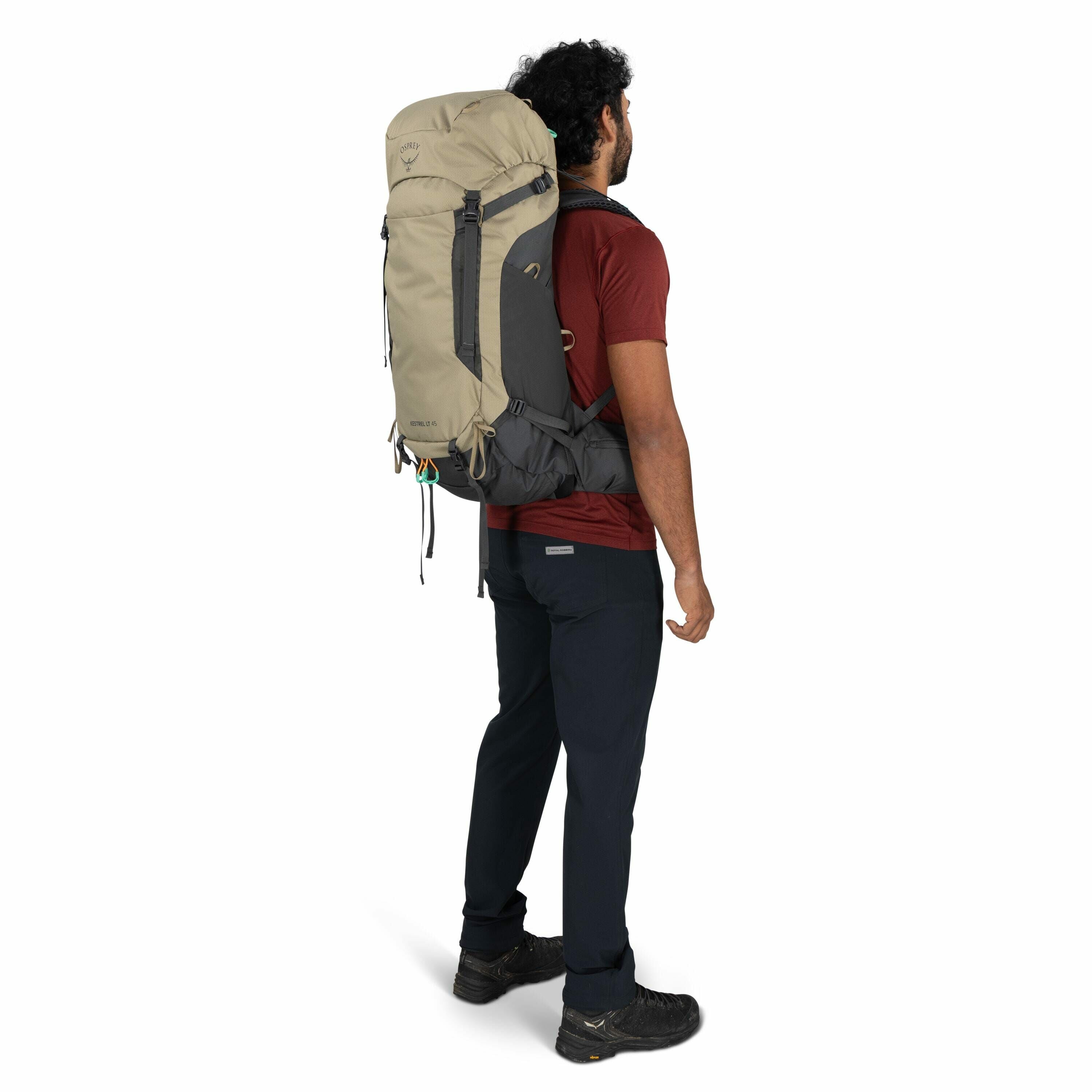 The image shows a man with dark curly hair standing sideways, wearing a large hiking backpack. The backpack is a combination of beige and dark gray colors with the Osprey logo visible on the top front, and the model name "Renn 65" is printed on the lower