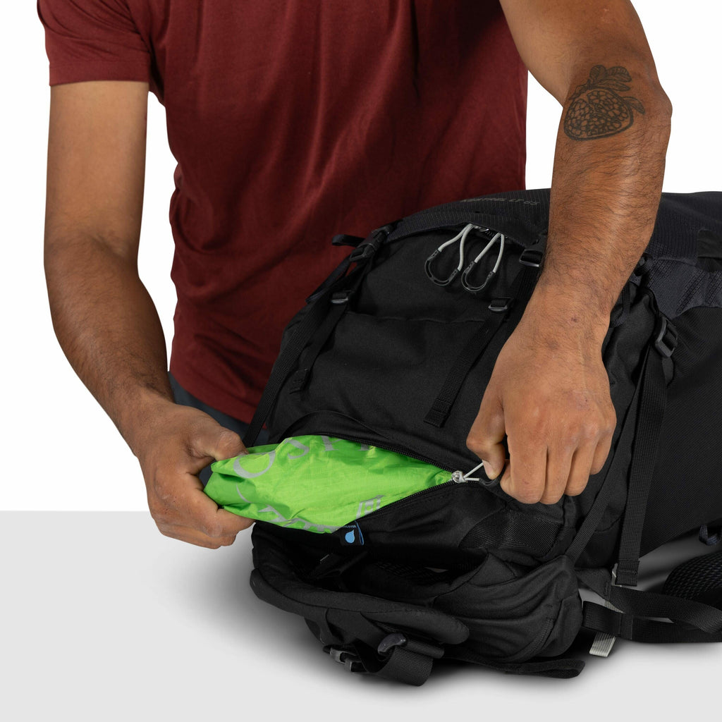 The image shows a person wearing a red shirt, zipping up a black backpack with their right hand while holding a green fabric in the other hand. The fabric appears to have blue and light green designs or text printed on it. The person's left arm is partial