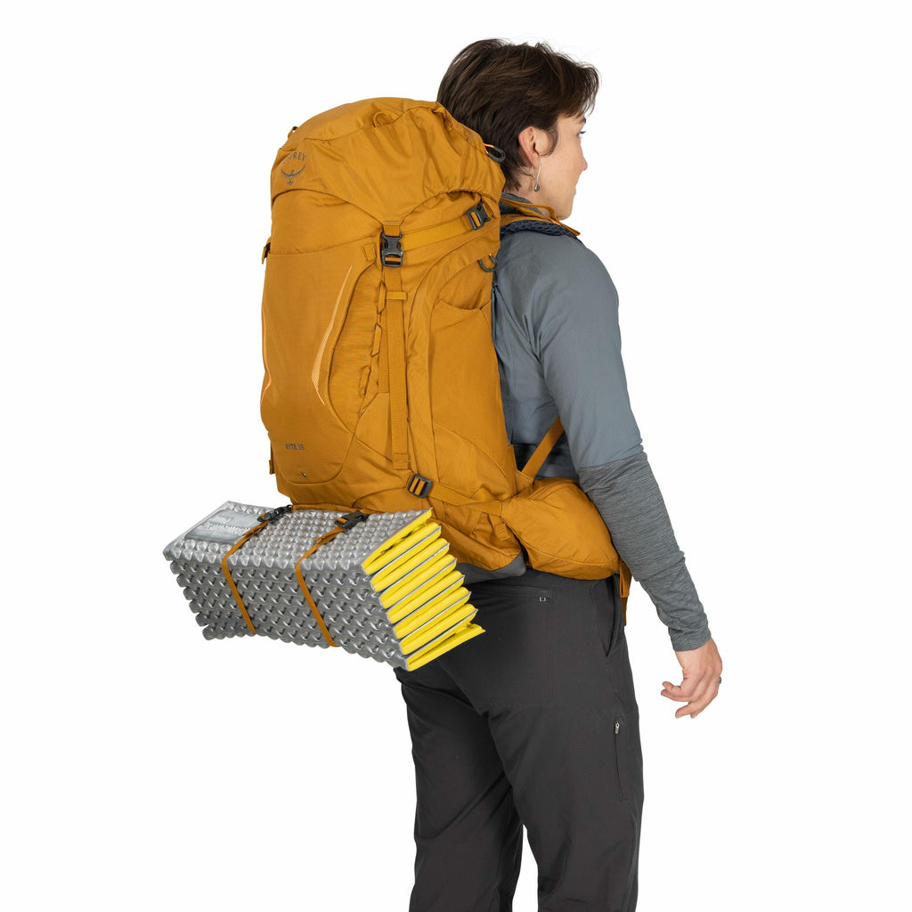The image features a person standing sideways wearing a large, mustard-colored Osprey brand hiking backpack. The backpack has various compartments and straps, including a visible buckle near the center. Attached to the bottom of the backpack is a folded,