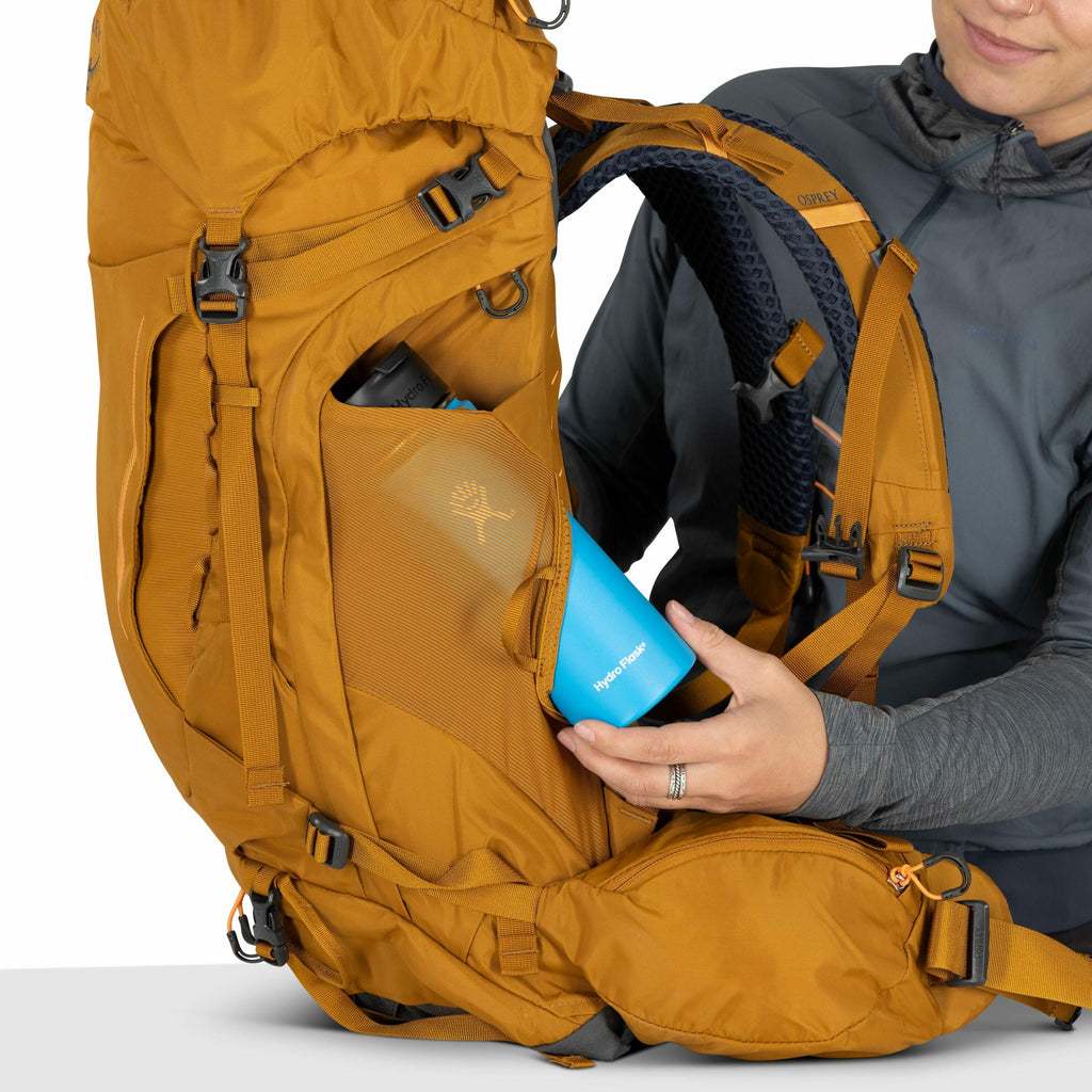 The image shows a person placing a blue Hydro Flask water bottle into the side mesh pocket of a mustard-colored Osprey backpack. The backpack is designed for outdoor activities and features adjustable straps and several compartments, including a hip belt