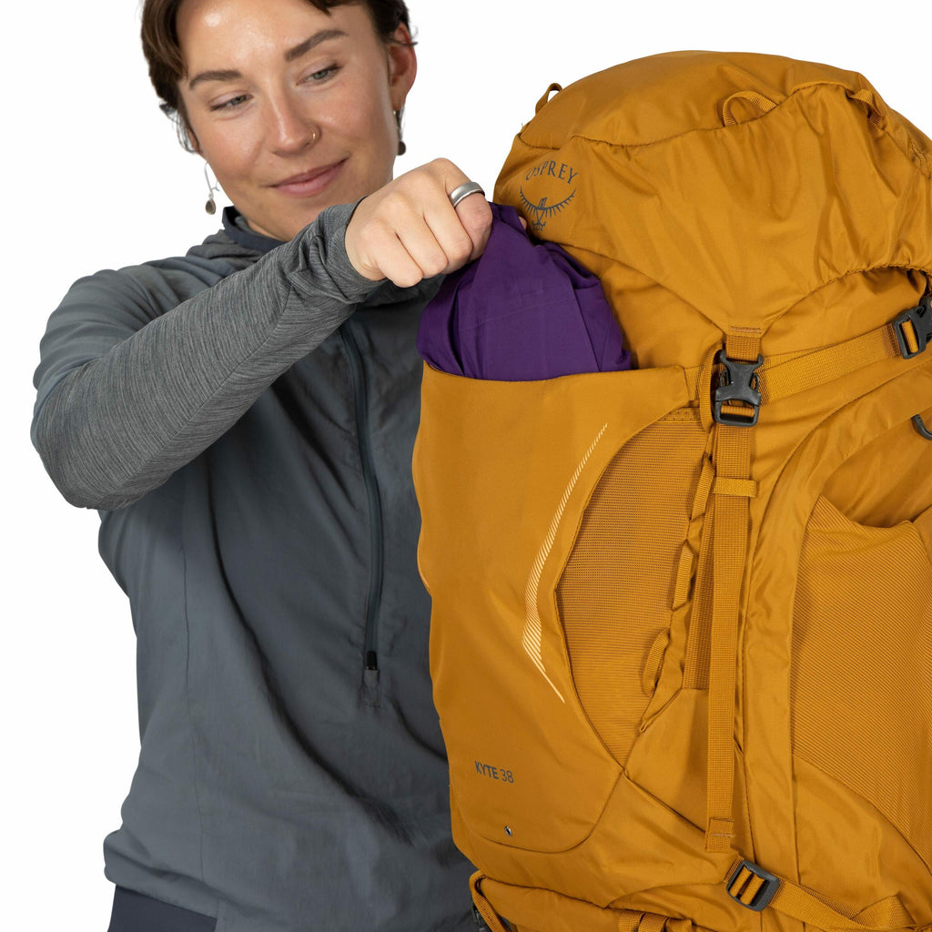 The image shows a person holding an orange backpack by Osprey with the model name "Kyte 38." The backpack is designed for hiking and has multiple compartments, including an accessible side pocket where the person is placing or retrieving a purple item. Th