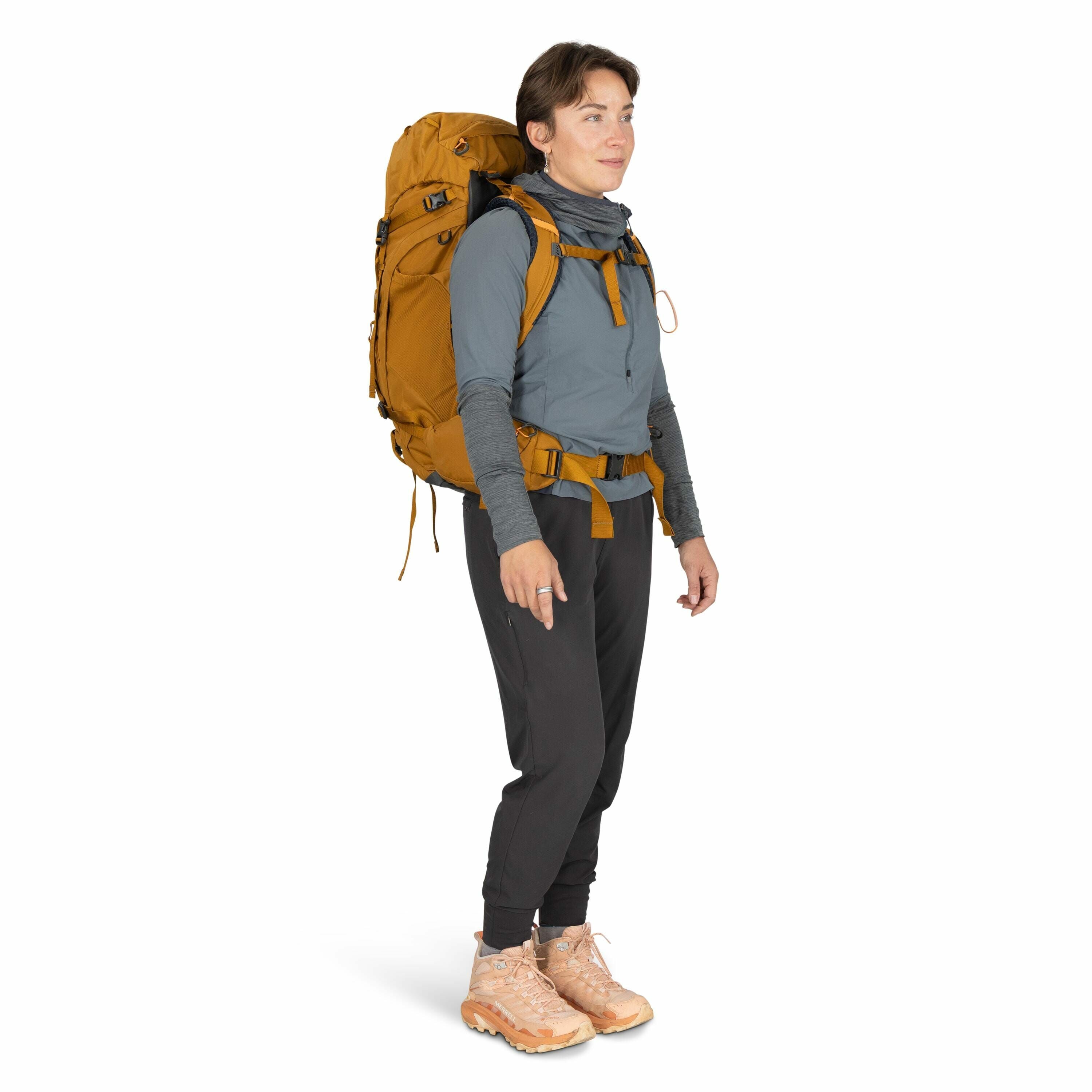 The image features a person wearing outdoor gear suitable for hiking or backpacking. The individual is dressed in a light blue long-sleeve shirt with a high-neck collar, layered over a grey shirt, and black athletic pants. They are carrying a large mustar