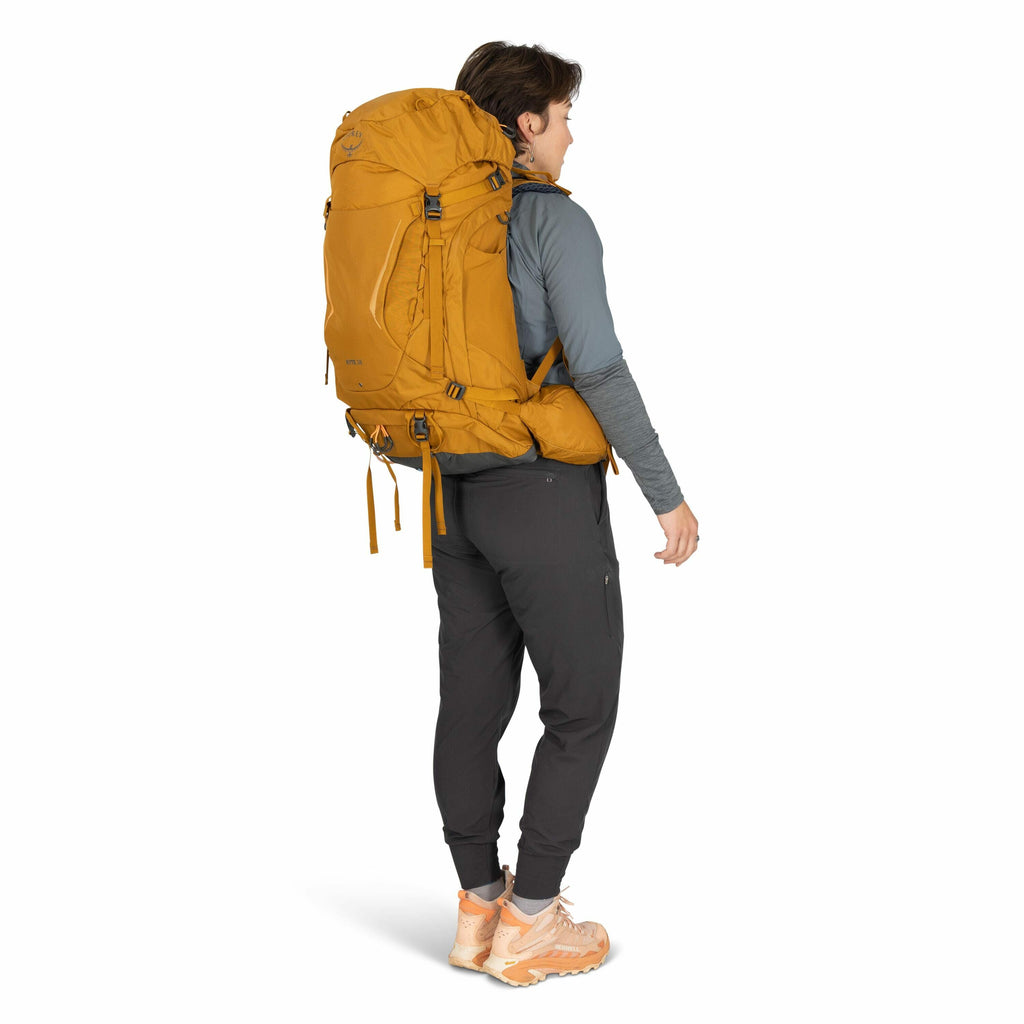 The image shows a person wearing outdoor gear, including a large, mustard-colored backpack branded with the Osprey logo and marked "Archeon 65." The individual is dressed in a long-sleeved gray top and dark pants, which appear to be suitable for hiking or